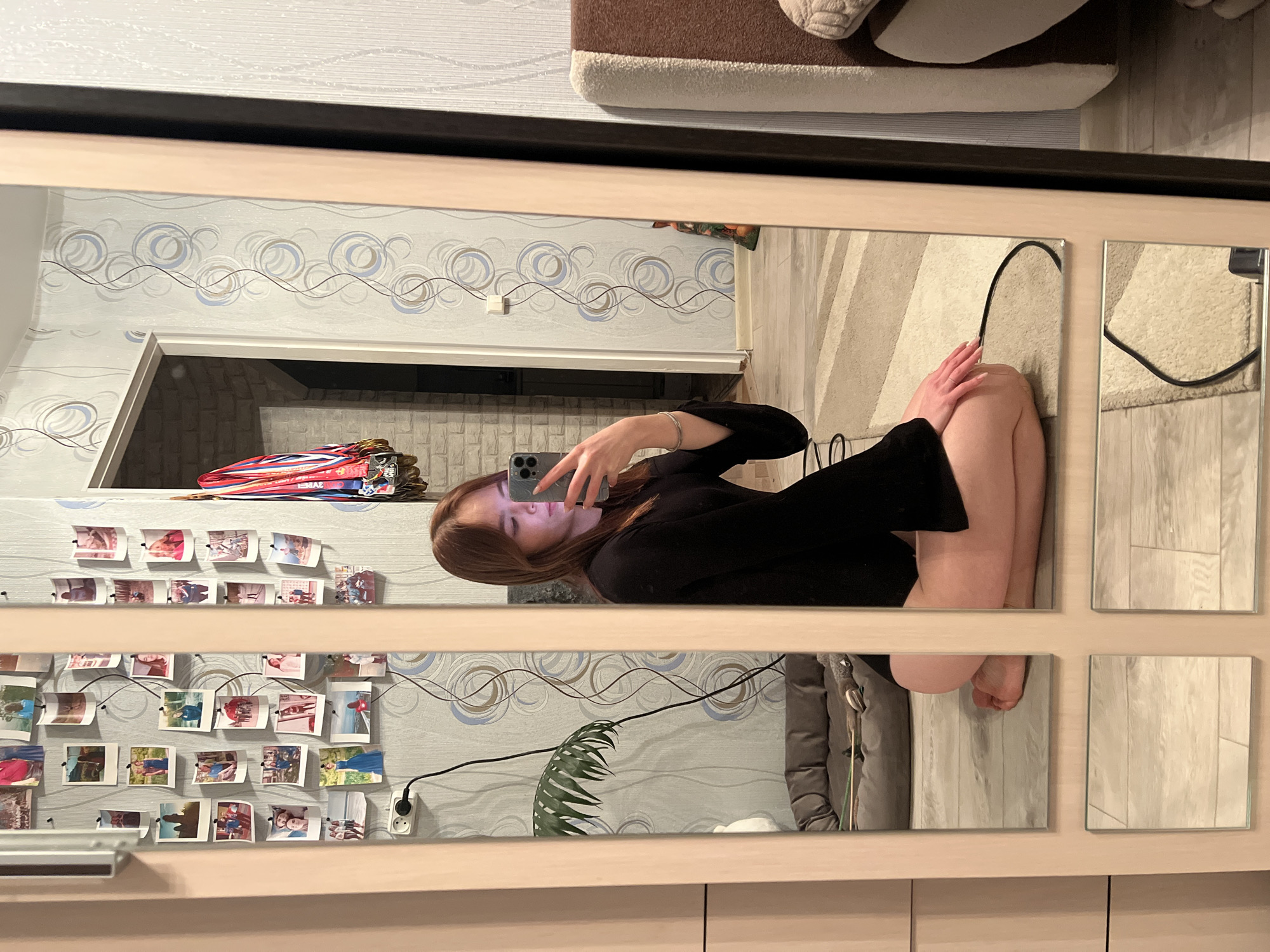 An interior scene captured as a mirror reflection depicts a young female in a residential dwelling in Chaikovsky, Russia. The female, with reddish-brown hair and wearing a black long-sleeved top, is kneeling on a light-colored floor, holding a light-colored smartphone with multiple cameras to take a selfie. A silver bracelet is visible on her left wrist.
The mirror, framed in light wood-toned material, reflects the room behind her. The wall to the left features light-colored wallpaper with a blue and grey concentric circle pattern, adorned with numerous small instant photographs depicting various individuals and scenes. A white electrical outlet with a black cord plugged into it is mounted on this wall. Below the outlet, a dark green leafy plant and a grey pet bed containing toys are visible.
To the right, a doorway leads into another area with a light-colored brick-patterned wall, from which several medals on colorful ribbons are suspended. The floor is light-toned laminate or wood, partially covered by a light-colored rug with subtle striped patterns. A portion of a sofa or bed with light brown and white upholstery is visible on the far right edge of the reflection.