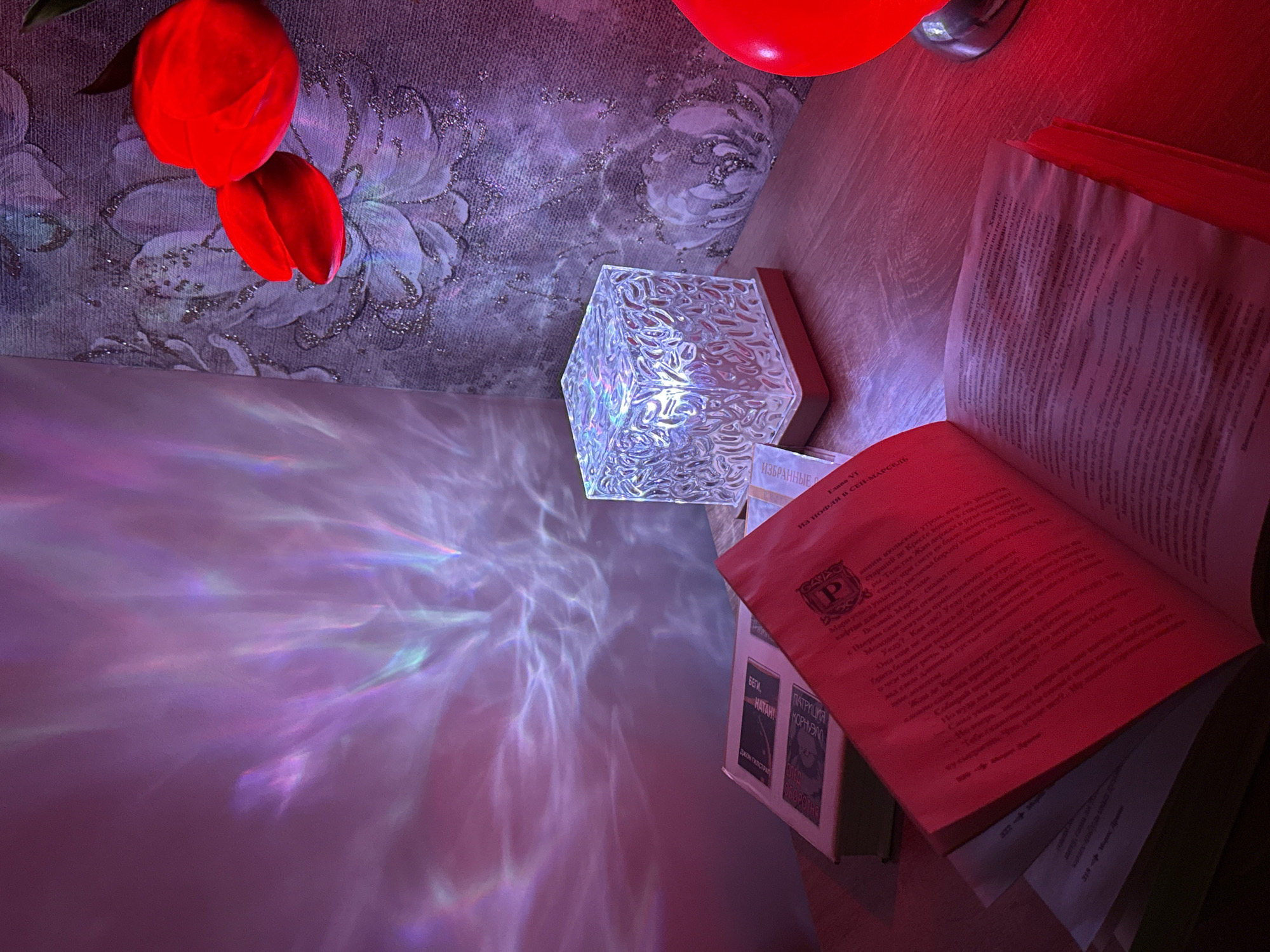 The image depicts an indoor scene, likely a reading nook or bedside table, set at night or in a dimly lit environment. The primary subjects include two bright red artificial tulips hanging against a textured wall, a distinctive cubic light fixture, an open book, and a small stack of books.
The wall behind the tulips features a purple-toned wallpaper with a floral pattern and glittering accents. The cubic light fixture, made of clear textured material, casts intricate, wavy patterns of purple, blue, and white light onto the adjacent wall and table surface, creating a dynamic visual effect. A red glow bathes the open book and the surrounding wooden surface. Partially visible in the top right is another red, possibly decorative, object.
The scene suggests a quiet moment of reading. The open book displays Cyrillic text, with "Глава VI" (Chapter VI) and "ИЗ НОФЛЯ В СЕН-МАРСЕЛЬ" (FROM NOFL TO SAINT-MARCEL) visible as a chapter title. A stylized 'P' logo is also present on the page. The text within the book appears to be narrative prose, including a phrase that translates to "I'll leave? How so? I'll leave this morning?"
The stack of books beside the open one also features Cyrillic titles and names, including "ИЗБРАННЫЕ" (SELECTED), "БЕГИ," (RUN,), "НАТАН!" (NATHAN!), "ПАТРУЛИЯ" (PATROL), "КОРНУЭЛ" (CORNWELL), "ДЖОН ГИЛСТРАЛ" (JOHN GILSTRAP), and "ОБОРОТНЯ" (WEREWOLF). The combination of artificial light, books, and decorative elements creates a cozy and contemplative atmosphere.