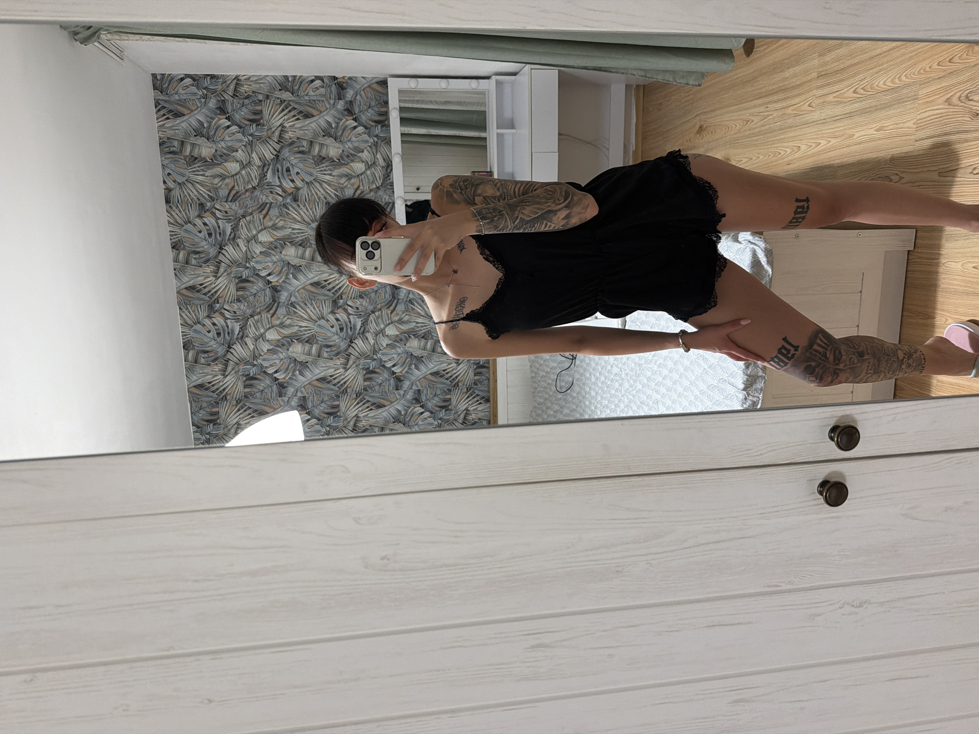 A young woman with dark hair and tattoos on her arms and legs stands in a bedroom, taking a mirror selfie. She is wearing a black romper. The room has a patterned wallpaper, a bed, and a vanity with lights. The image is taken from a slightly high angle, showing the woman from the knees up.
