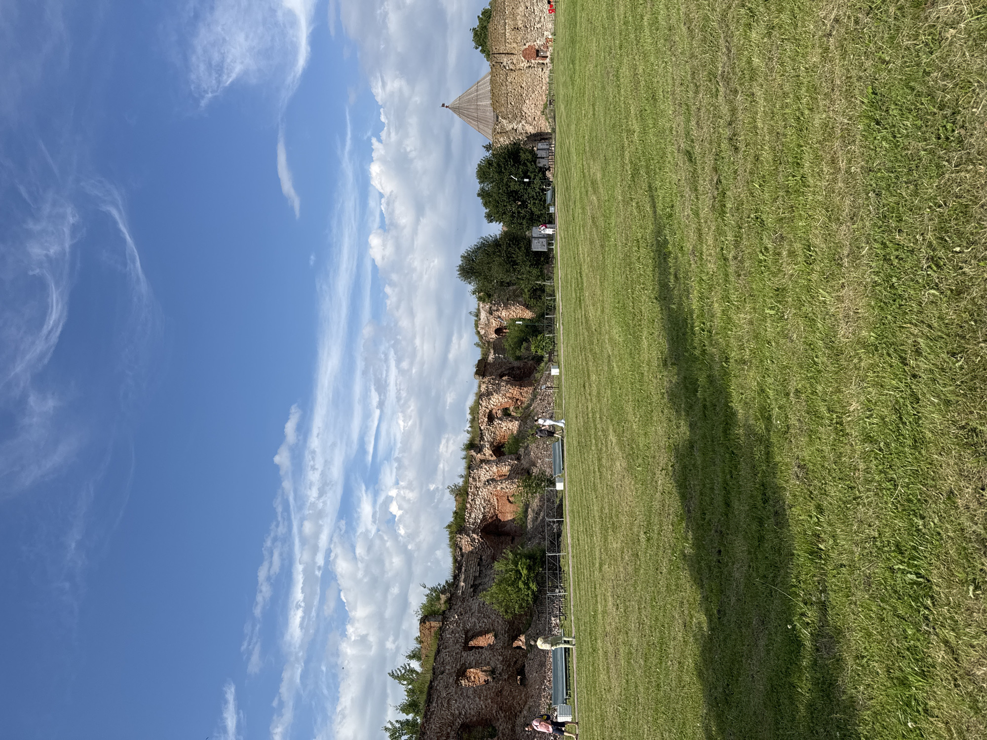 The image depicts a sunny day at the Oreshek Fortress in Shlisselburg, Russia, with a clear blue sky dotted with white clouds. In the foreground, a vast expanse of well-maintained green grass stretches towards ancient stone fortifications. The fortress walls, constructed from weathered red bricks, show signs of age and decay, with visible arches and openings. Several people are visible walking on a path in front of the fortress, and some benches are placed along the path. To the right, a conical wooden structure, likely a tower or roof, stands atop a section of the stone wall. Lush green trees are scattered around the fortress grounds, adding to the picturesque scenery. The overall impression is one of historical significance and natural beauty.