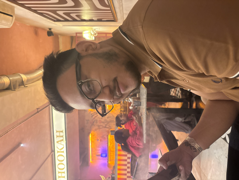 A man wearing glasses and a tan polo shirt stands in what appears to be a hookah lounge. The lounge has orange walls, and a neon sign says "HOOKAH." Two people are visible in the background, seated at a table. Vapor or smoke is prominent in the lower center of the frame. The man has a bracelet on his left wrist and is holding a dark object. A patterned ceiling and an air conditioning unit are also visible.