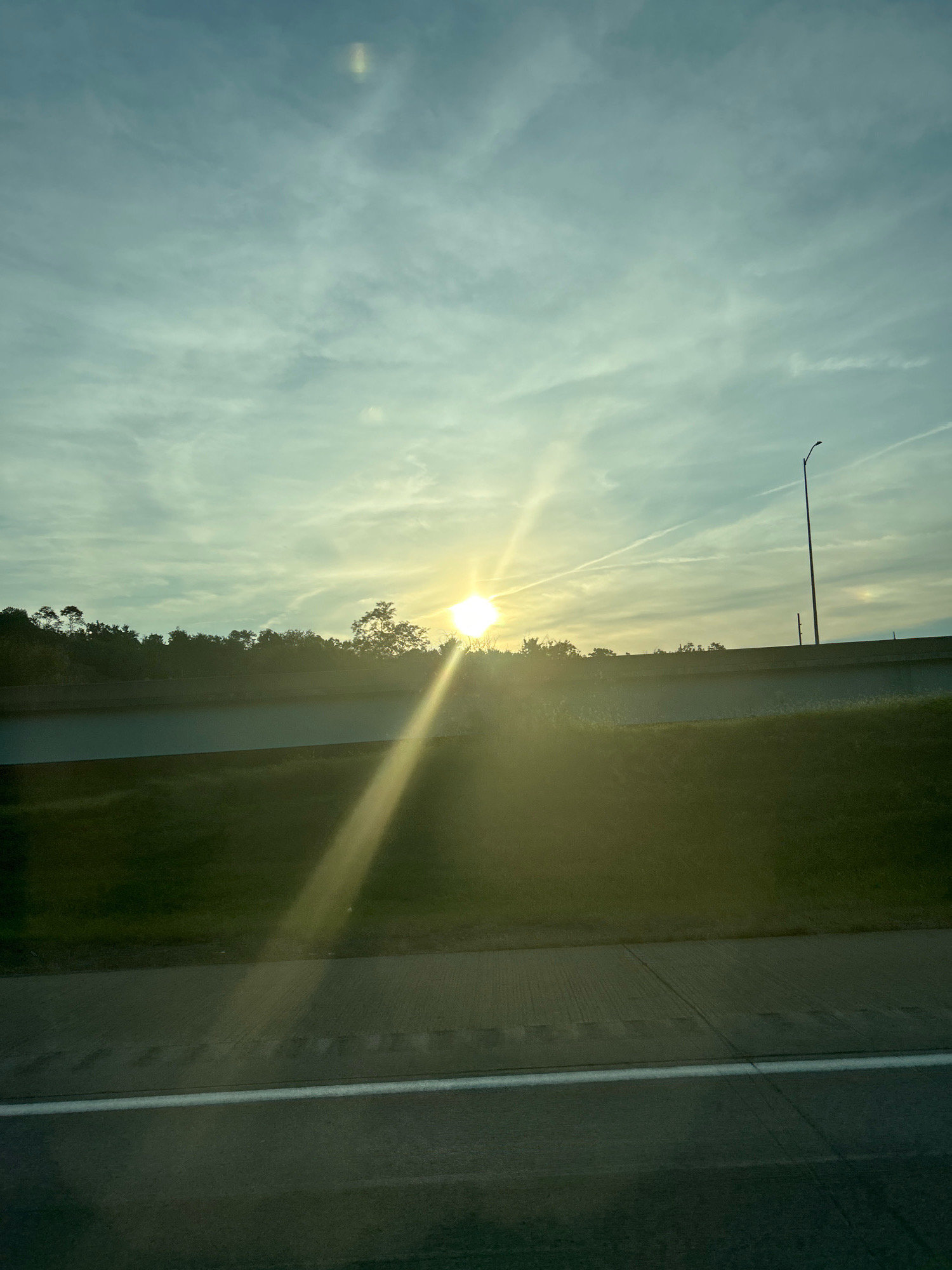 A bright sun shines through a cloudy sky, casting a long light ray across the image. A line of trees is silhouetted against the sky, with a highway overpass in the middle ground. A street light stands tall on the right side of the overpass. The foreground shows a grassy slope and a portion of a road. The scene is likely a roadside view of a rural or suburban area.
