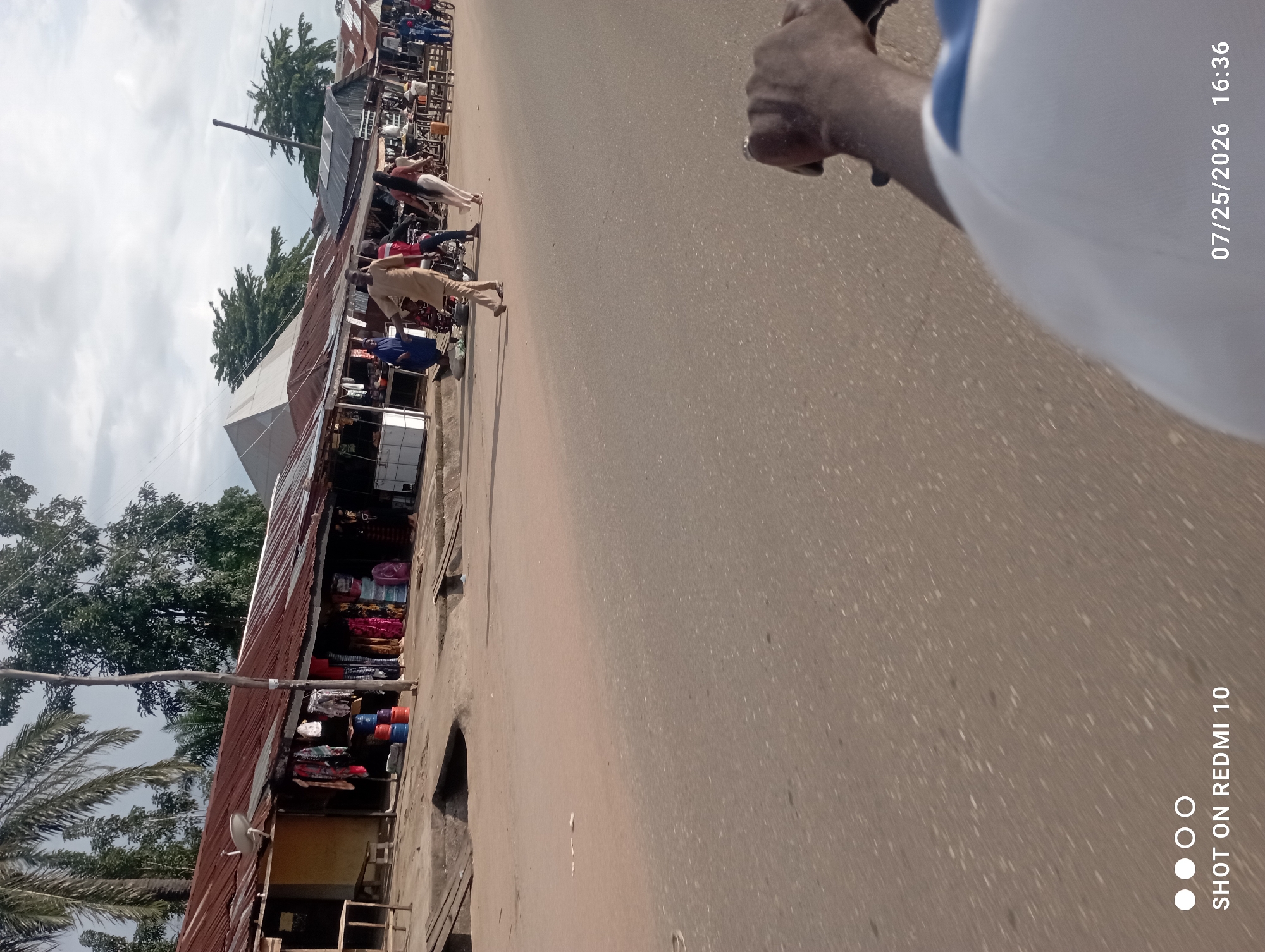 The image shows a street scene in Serti, Nigeria, with a row of market stalls lining one side of the road. The stalls have corrugated metal roofs and appear to be selling clothing and other goods. Several people are visible walking and interacting in the market area, including a person on a motorcycle. The road is made of asphalt, and there is a concrete sidewalk in front of the market. The sky is cloudy. The image was taken on July 25, 2026, at 16:36.