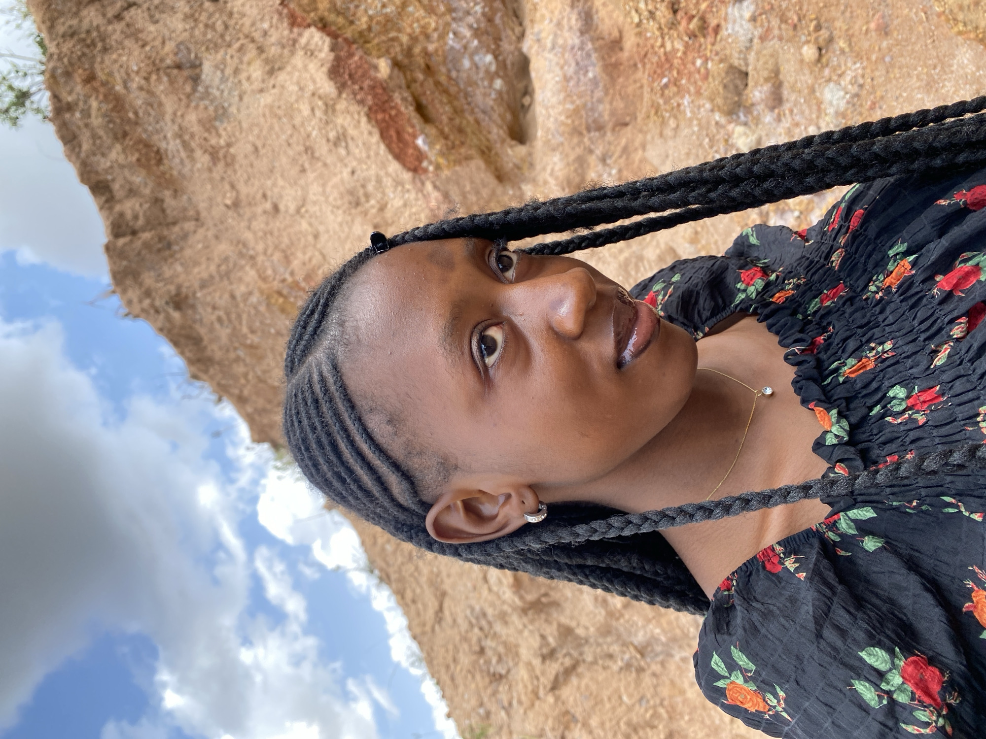 A young Black woman with long, braided hair and a floral black top is shown from the chest up, looking to her right. She is wearing a silver hoop earring and a delicate gold necklace with a single diamond pendant. The background consists of a rocky, orange-hued hillside under a cloudy blue sky. This scene is located in Gura Top, Nigeria.