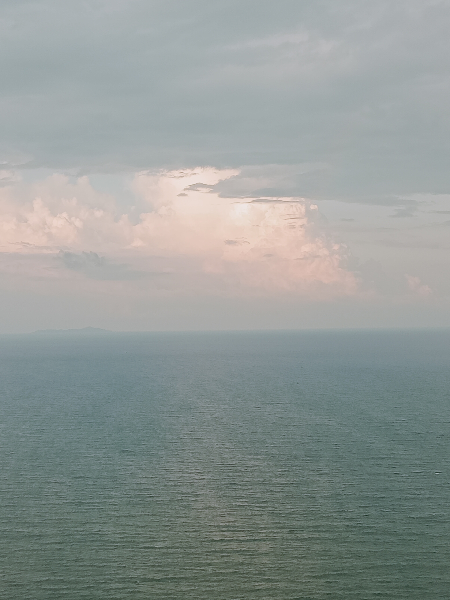The image depicts a seascape under a cloudy sky. A distant island is visible near the horizon. The water is a muted teal color with subtle wave patterns, and the sky is a mix of pale grey and pink hues, particularly around the cloud formations. The photo appears to be taken from an elevated position, overlooking the ocean. Without additional context, it's difficult to determine the precise location within the city of Nha Trang Ward, Vietnam, but the scene captures a view of the ocean and sky.