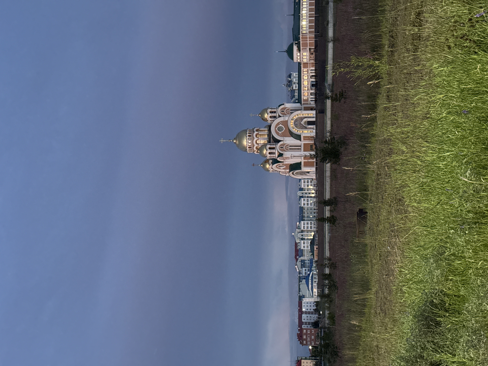 The image captures a view of the Sacred Cathedral of the Assumption of the Blessed Virgin Mary in Salekhard, Russia, at twilight. The cathedral, with its prominent golden domes and intricate architectural details, is illuminated, standing out against the darkening sky. Behind the cathedral, a row of modern, multi-story residential buildings with lit windows stretches across the horizon, suggesting an urban setting. In the foreground, a field of tall, dry grass and some green foliage creates a natural boundary between the viewer and the constructed environment. The overall atmosphere is serene, with the muted colors of the sky and the warm glow of the buildings and cathedral contributing to a peaceful, evening ambiance. There are no people or animals visible in the image. The scene appears to be a static, observational shot, focusing on the landmark and its surroundings.