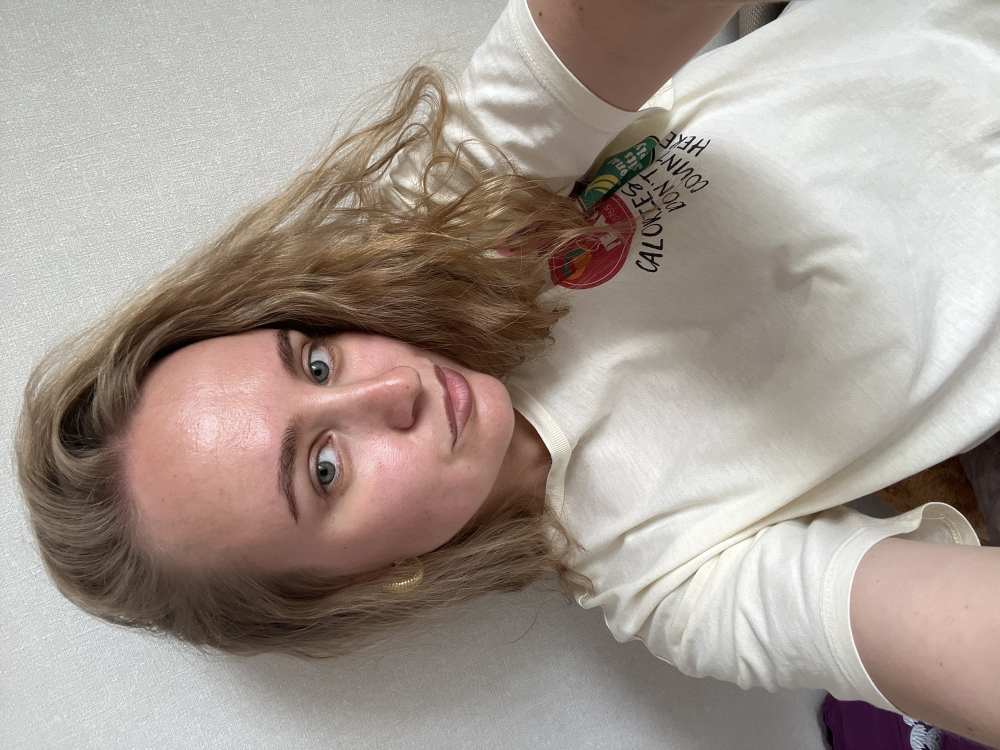 A young woman with blonde hair and blue eyes, wearing a cream-colored t-shirt with a graphic print, takes a selfie against a textured white wall. The t-shirt has a circular graphic with text that reads "CALORIES DON'T COUNT HERE." She is wearing a gold hoop earring. The scene appears to be indoors, likely a bedroom or living area, based on the soft lighting and the plain wall. No specific landmarks or contextual cues are present to indicate the city of Chaikovsky, Russia, or any urban environment.
