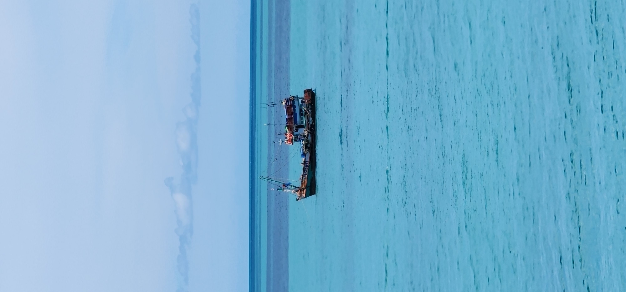 A fishing boat is anchored in the turquoise waters off the coast of Baan Chaweng Noi, Thailand. The sky above is a pale blue with scattered white clouds. The ocean is calm, with gentle ripples visible on the surface. The fishing boat is painted in various colors, including green, blue, and orange, and has several masts and fishing equipment. There are no people visible on the boat. The scene is tranquil and serene, conveying a sense of peace and quiet.