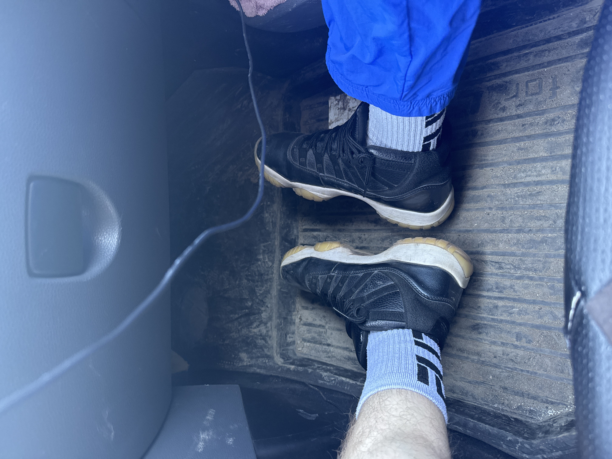 A close-up shot shows a person's feet wearing black sneakers with white soles and light grey socks with black stripes. The sneakers are positioned on a dark, textured floor mat with the word "TOR" partially visible, suggesting the interior of a vehicle. A light grey surface is seen on the left side of the frame. A blue object is visible near the top. The location is not possible to deduce.