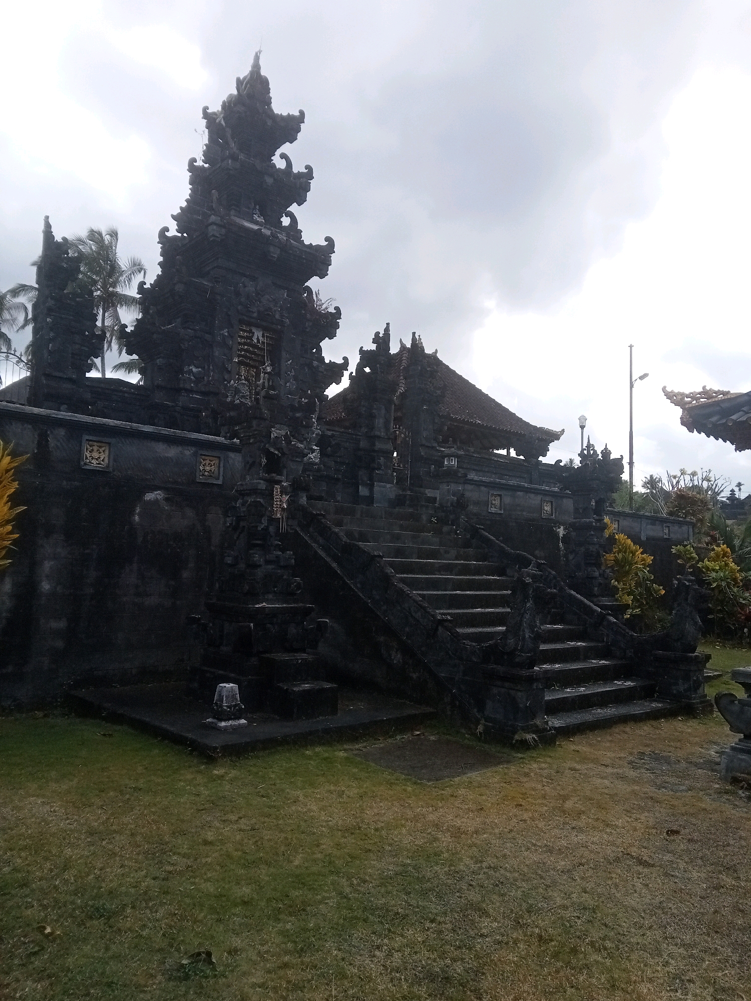 The image shows a stone temple structure with intricate carvings and a tiered roof, set against a cloudy sky. In the foreground, a grassy lawn is visible with some dry patches and a small stone monument. A set of stone stairs leads up to the temple entrance, flanked by ornate railings. Palm trees are visible in the background to the left of the temple. A streetlamp is also present in the background to the right. The overall scene appears to be in a tropical or subtropical region given the presence of palm trees.