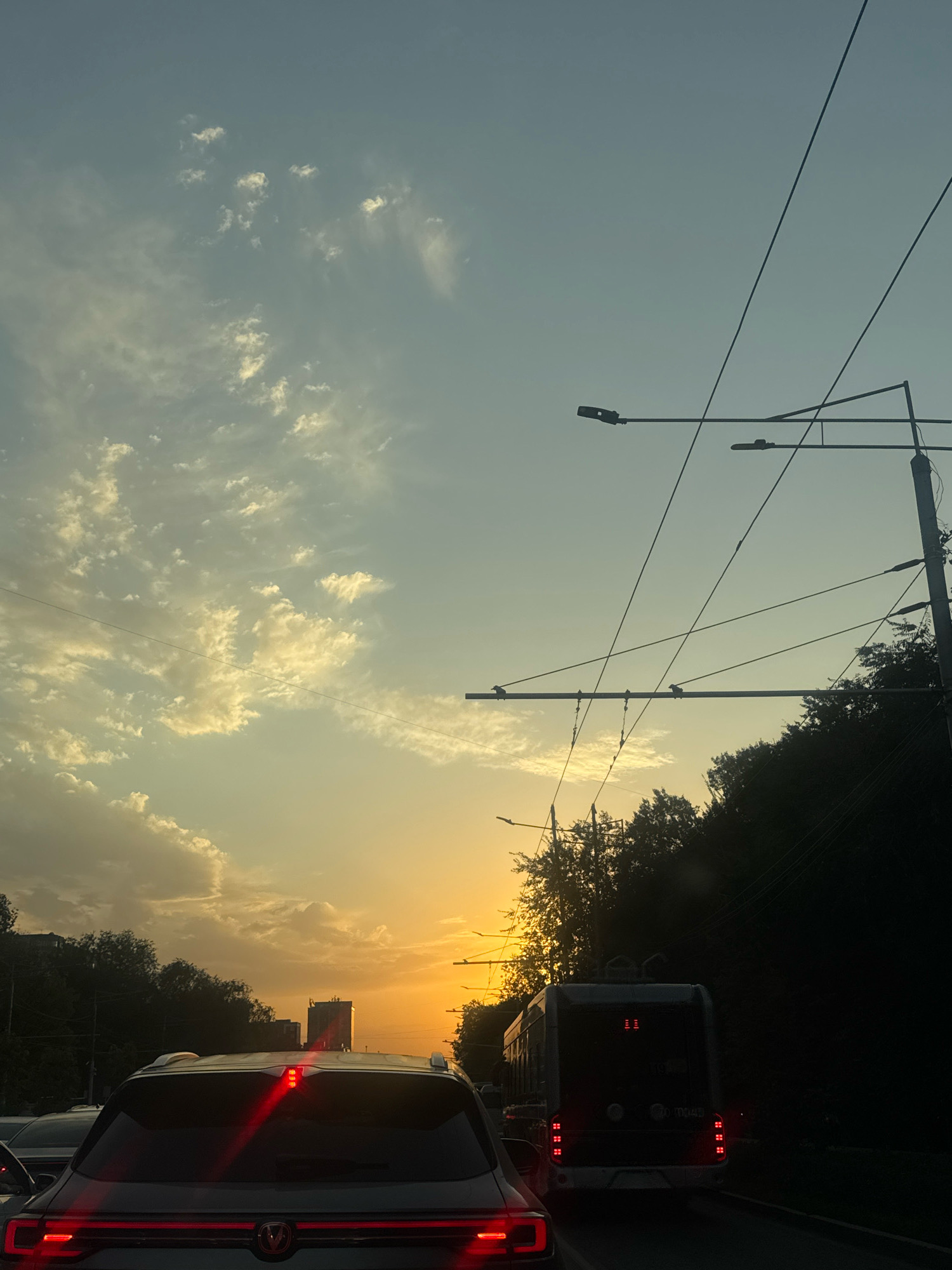 The image captures a sunset view from a vehicle in traffic, likely on a street in Almaty, Kazakhstan, given the presence of trolleybus wires. The sky is a gradient of pale blue near the top, transitioning to hues of yellow and orange as it approaches the horizon. Scattered, wispy clouds are illuminated by the setting sun.
In the foreground, the rear of a dark-colored SUV is visible, with its brake lights illuminated red. To the right and further down the road, a trolleybus is stopped in traffic, its red taillights also lit. The trolleybus has the number "7096" visible on its rear. Overhead, multiple trolleybus wires and streetlights are silhouetted against the sky.
The scene is bordered by dark trees on the right, obscuring the background on that side. In the distance, on the left side of the frame, a tall building with a dark facade stands against the sunset. The overall impression is of a city street experiencing congestion during the golden hour.