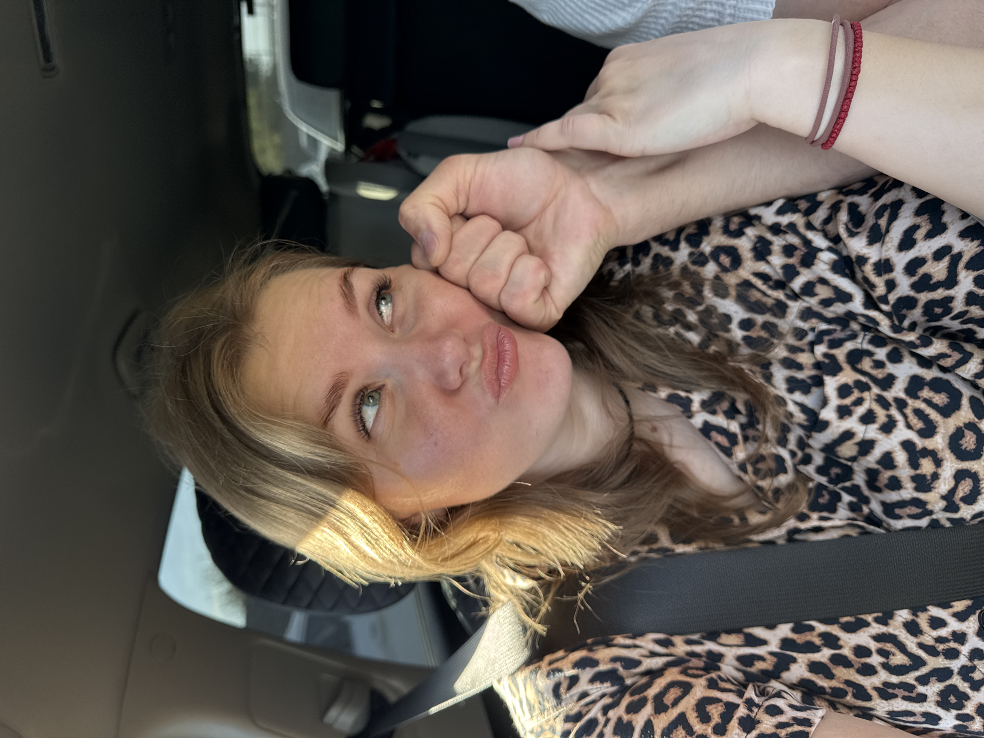 A young woman with blonde hair and green eyes is posing in a car. She is wearing a leopard print shirt and a seatbelt. Her left hand is on her cheek, while another person's hands are above her hand. The person on the top hand is wearing a red bracelet. The image appears to be taken inside a car. There is no indication that the image was taken in the city of Perm, Russia.