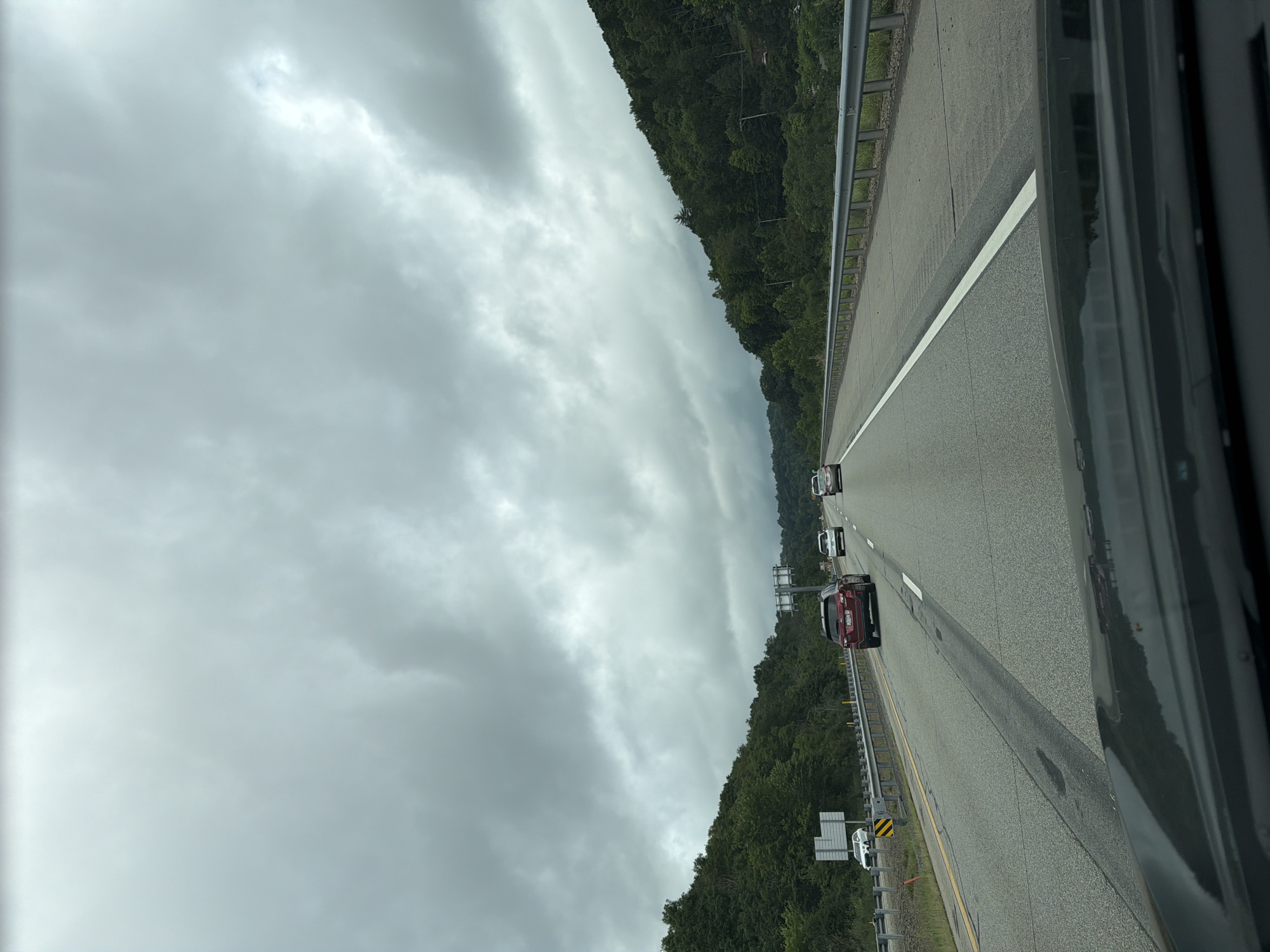 An overcast day on a multi-lane highway in the vicinity of Mount Nebo, USA. The road is paved with visible lane markings and guardrails along the right side. Dense, green trees line the hilly landscape on both sides of the highway. Several vehicles are driving on the road, including a dark red SUV in the right lane and a white car in the left lane further ahead. The sky is heavily clouded, creating a dim and diffused light.