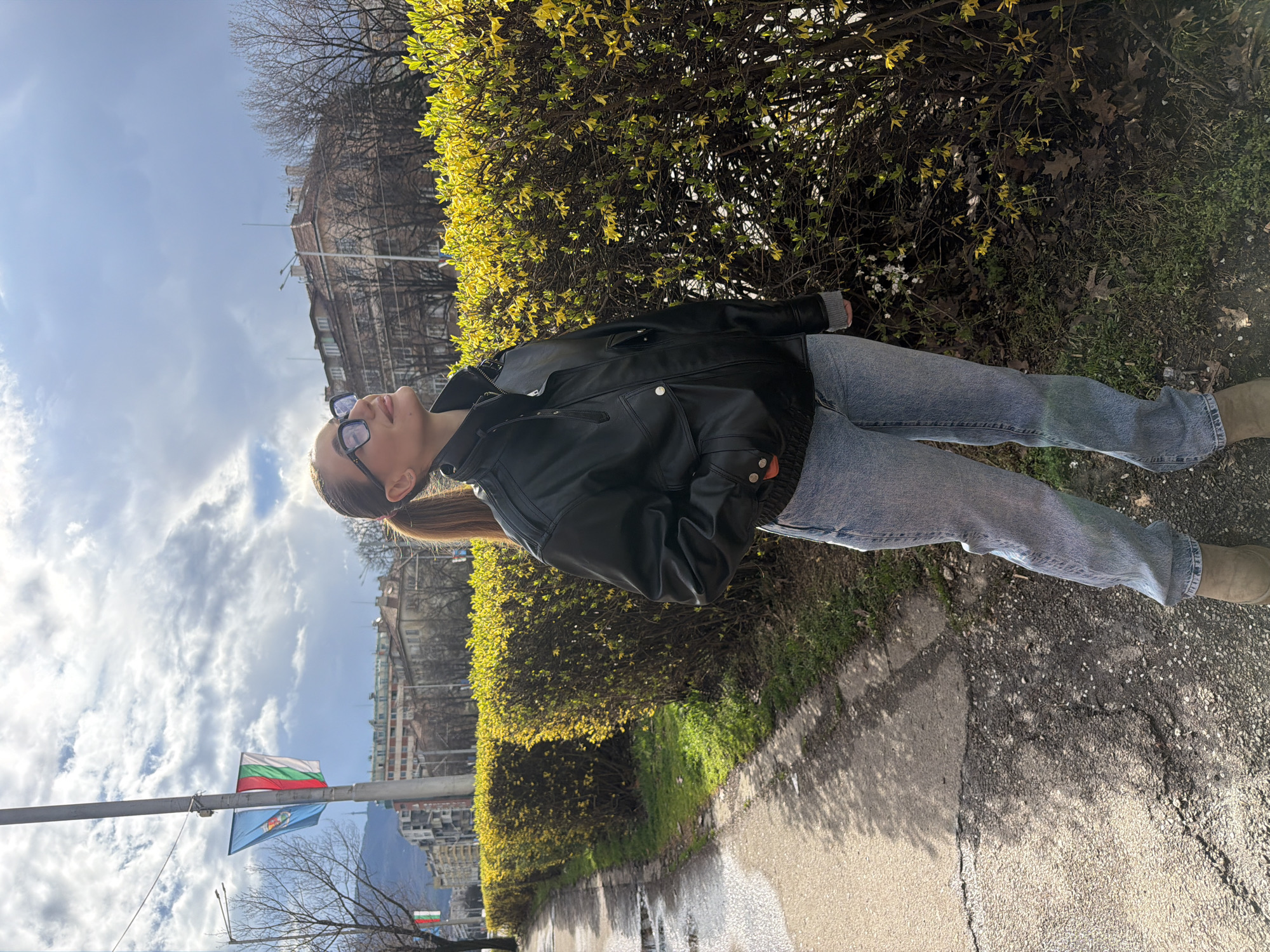 A woman is lying on her side next to a trimmed bush on a sidewalk. She is wearing a black leather jacket, light blue jeans, and brown boots. She has glasses on and her hair is in a ponytail. She is looking up. The sidewalk is wet. In the background, there are buildings, a Bulgarian flag, and a flag of Sofia near the pole. The photo was taken in Sofia, Bulgaria.