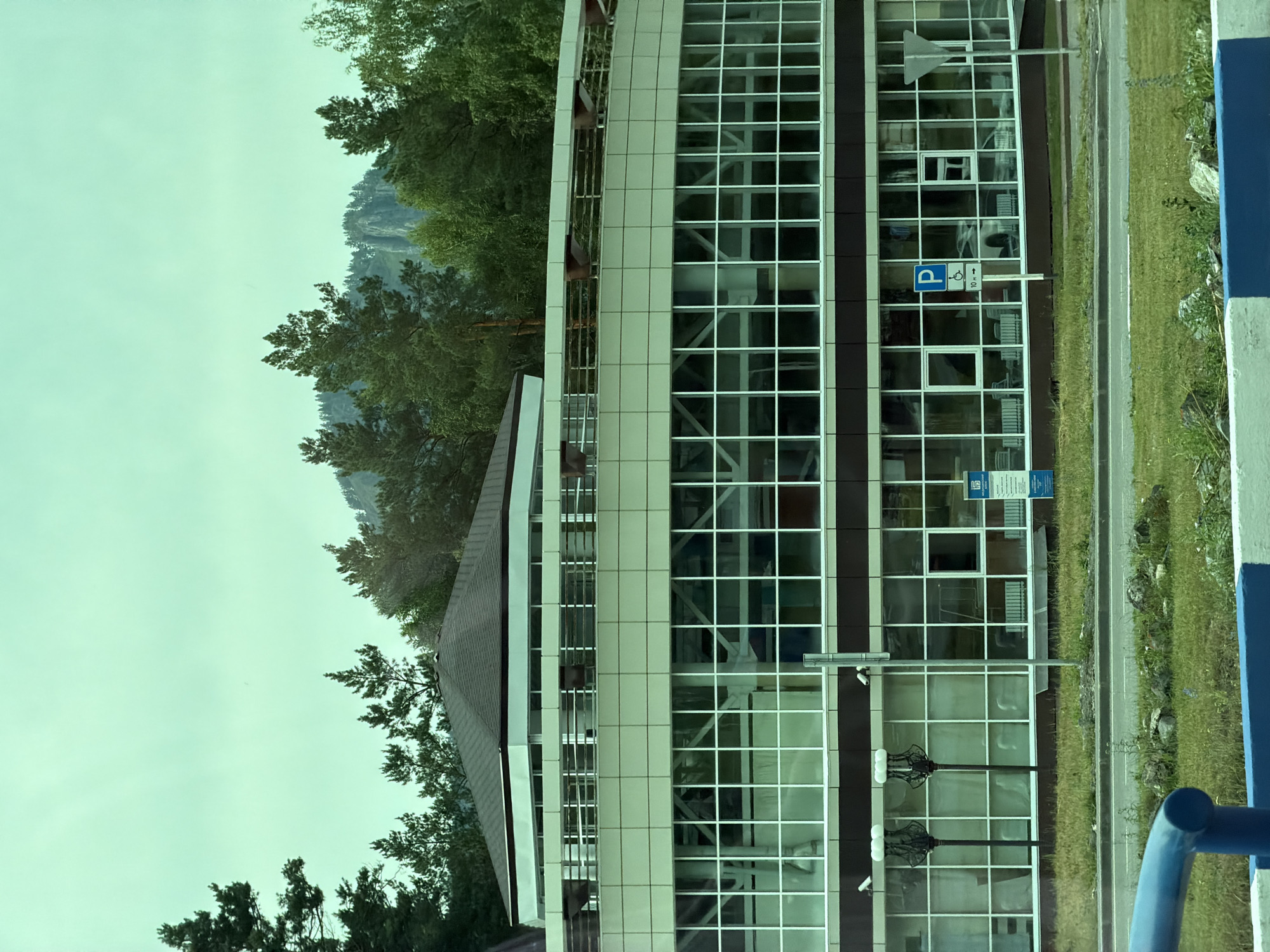 The image shows a modern building with a glass facade, situated in a natural landscape with trees and mountains in the background. The building appears to be multi-story with a grid-like pattern of windows. There are trees on the left side of the building, and a grassy area with a road next to it on the right. A blue and white striped barrier is visible on the far right.
On the building's facade, there are signs. One sign is a blue square with a white "P" symbol and an accessibility icon, indicating a parking area. Another sign above it is triangular and white. There are also two streetlights with decorative tops visible on the ground floor level. Security cameras are also mounted on the building.
The sky is overcast, suggesting a cloudy or hazy day. The natural setting implies the building is located in a scenic or somewhat remote area. There are no people visible in the image. The scene is static, with no activity taking place. The overall impression is one of a functional modern structure integrated into a natural environment.