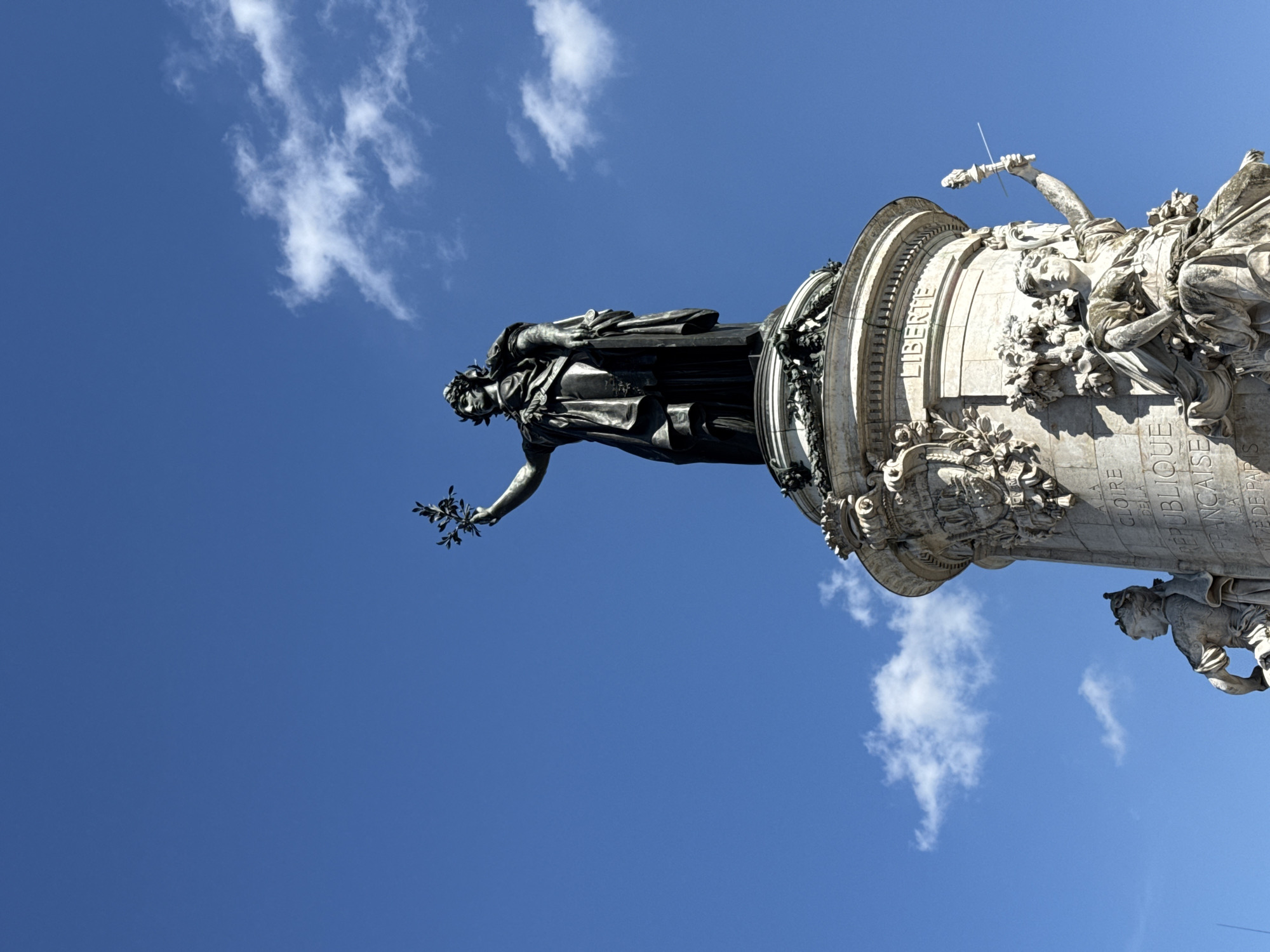 The image depicts the top portion of the Place de la République monument in Paris, France, against a blue sky with scattered white clouds. The bronze statue of Marianne, the symbol of France, is at the top of the column. "LIBERTE" is carved on the column. Underneath, there are reliefs and sculptures on the column.