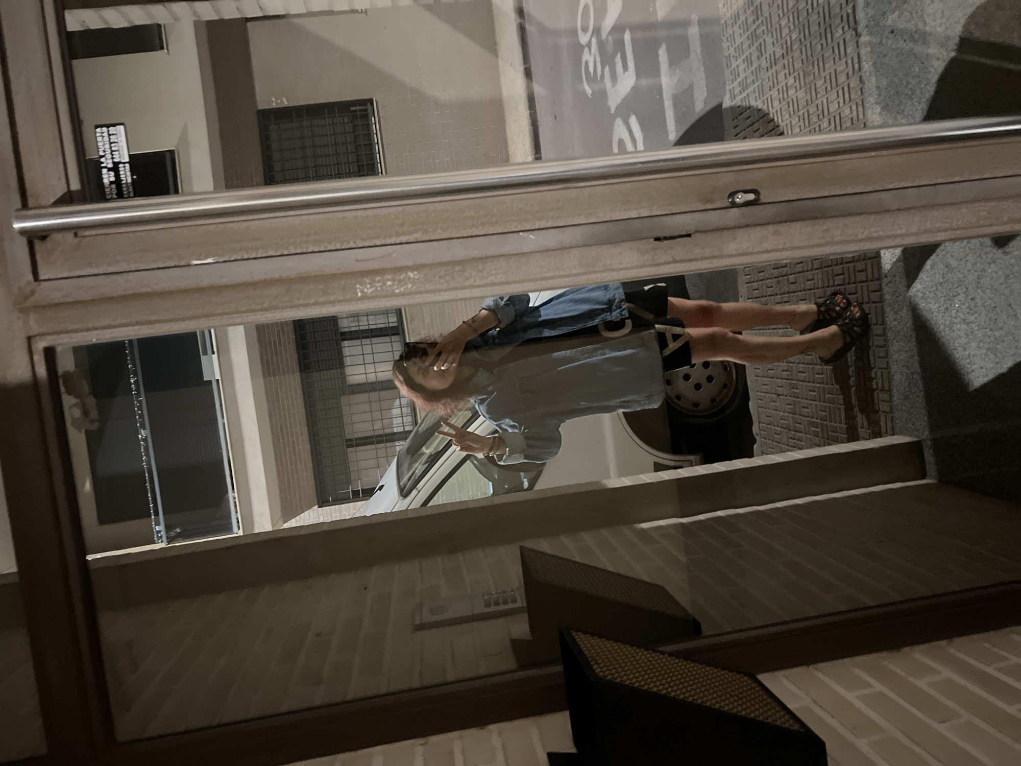 An individual is captured in a selfie taken in the reflection of a glass door at night. The female subject, positioned centrally, is holding a smartphone to her face, partially obscuring her features, and making a peace sign with her left hand. She has reddish-brown hair and is wearing a light blue denim jacket over a dark garment, possibly a dress or top with white markings. Her feet, clad in dark, possibly wedge sandals, are visible on a patterned tiled surface.
The reflection reveals the exterior environment beyond the glass door, which features a street and adjacent buildings under artificial lighting. A white van is parked on the street behind the subject. The asphalt displays painted white markings, including the numbers "2" and "30". The architecture visible in the reflection consists of multi-story buildings with light-colored brick facades and dark-grated or shuttered windows. A balcony with a metal railing is also present.
A black sign with white Spanish text is clearly reflected in the upper right, reading: "CIERREN LA PUERTA POR SU SEGURIDAD" (Close the door for your security), followed by "ALARMA VERISURE" and a phone number "964 83 83 80". The glass door itself has a prominent vertical metal handle and a visible keyhole. To the immediate left within the internal space, a brick wall and a dark, woven object (possibly a planter or furniture armrest) are reflected. The scene is located in the city of Picassent, Spain.