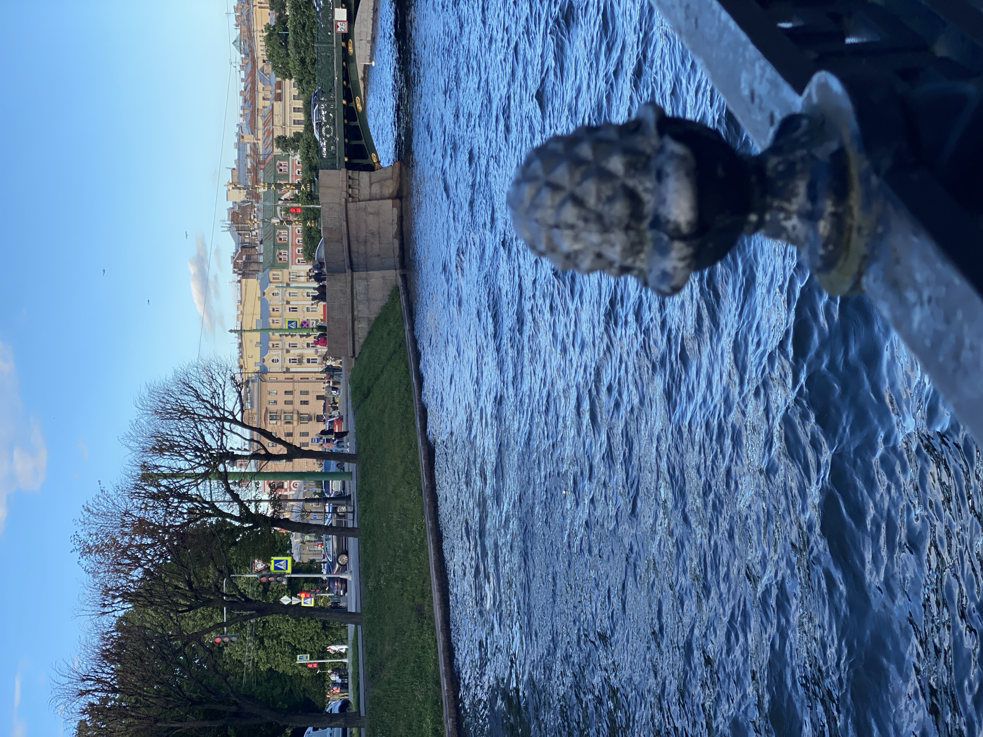 The image captures a view of the Fontanka River in Saint Petersburg, Russia, on a sunny day with a few clouds. In the foreground, a dark metal railing with an ornate finial runs along the edge of the riverbank. The river itself is a deep blue with choppy waves. Across the river, a stone bridge with an arched green metal structure spans the water. Beyond the bridge, the cityscape of Saint Petersburg unfolds, featuring buildings with distinct architectural styles, including those with mansard roofs and ornate detailing. A tree-lined embankment with a grassy slope is visible on the left side of the image, with a sidewalk and road running parallel to it. Several cars are parked and driving on the road, and pedestrians can be seen walking along the sidewalk and crossing the street. Traffic lights and pedestrian crossing signs are also present. In the distance, several small airplanes are visible in the bright blue sky.