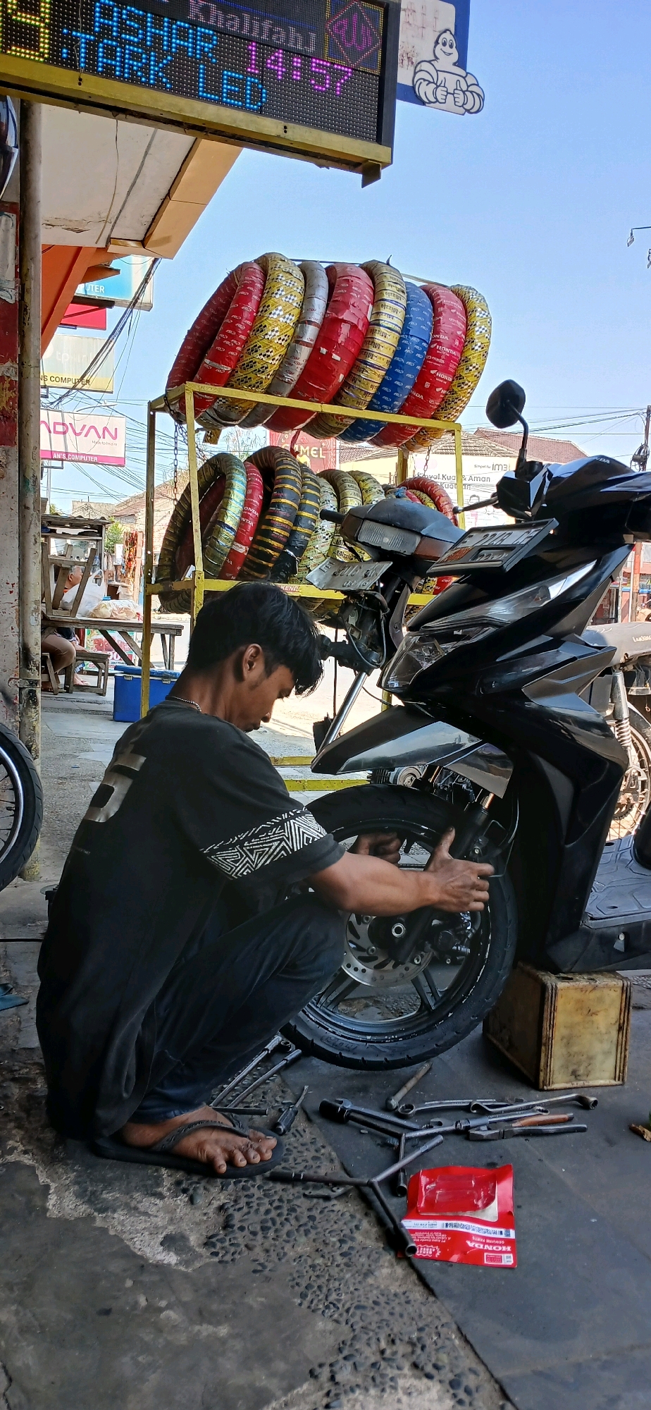 The image depicts a street-level mechanic's shop in Cirebon, Indonesia, likely on a sunny day given the bright lighting and clear blue sky. The shop appears to be open-air, with a roof structure providing shade.
The central figure is a young man, possibly in his early twenties, dressed in a black t-shirt with a decorative pattern on the sleeves and black pants. He is crouched down, actively working on the front tire of a black motorcycle. His hands are engaged with the wheel, suggesting he is either changing or repairing it. Tools, including wrenches and a lug wrench, are scattered on the ground around him, along with a red package of Honda genuine parts.
Behind the mechanic, a prominent yellow metal rack displays a colorful assortment of new motorcycle tires, stacked neatly in rows. The tires vary in color, featuring red, yellow, blue, and white packaging, indicating different brands or types. A motorcycle is parked next to the rack, partially obscuring it.
In the background, there are glimpses of other elements of the street scene. A sign with blue and pink LED lettering, displaying what appears to be information or advertisements, is visible above the tires. Further in the background, other shops or buildings can be seen, along with a person sitting at a table under a shaded area, possibly a customer or another vendor. The overall environment suggests a typical busy street commerce setting in an Indonesian city.