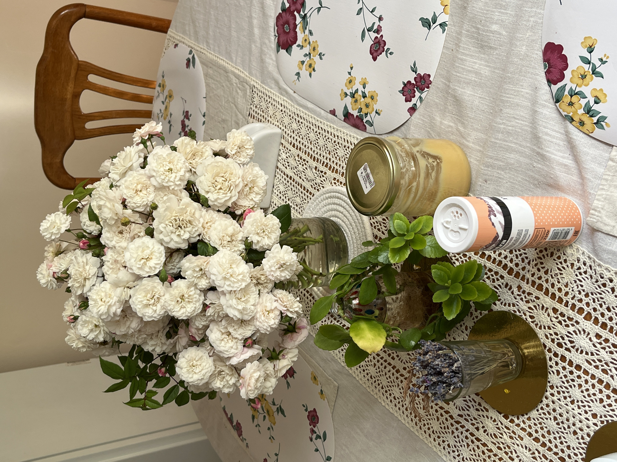 A floral-themed table is set with a bouquet of white roses in a glass vase, a small glass of dried lavender, a jar of honey, and a cylindrical container. The table is covered with a light-colored tablecloth and a crocheted runner. Two floral placemats are visible, one partially obscuring the other. A wooden chair is positioned behind the table. The lighting suggests an indoor setting, possibly a dining room, with natural light coming from the left. The overall scene conveys a domestic and somewhat rustic ambiance.