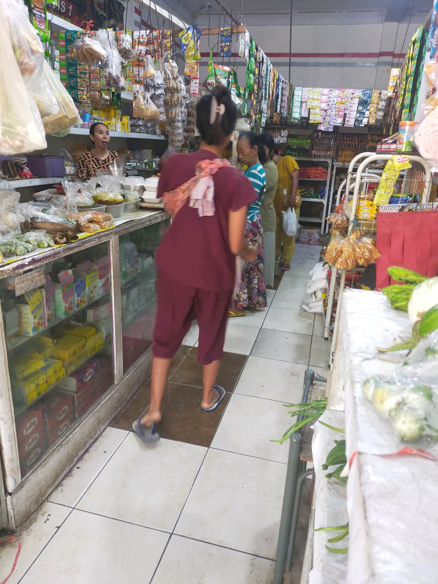 The image depicts the interior of a small convenience store in Mojodanu, Indonesia. The store is densely stocked with various goods, including bagged snacks, canned items, and packaged foods displayed on shelves and hanging from the ceiling. In the foreground, a woman wearing a maroon t-shirt and shorts is walking away from the camera, her back towards the viewer. She is wearing blue and white flip-flops. To her left, a glass display case is filled with boxes of packaged food items. Further back, three other individuals are visible. One woman, wearing a patterned black and beige top, is standing behind the counter, interacting with a customer. To her right, two other women are standing, one in a blue and white striped t-shirt and the other in a yellow t-shirt, both holding shopping bags. The store has a tiled floor with a checkerboard pattern of light and dark brown tiles. The environment appears to be a typical Indonesian neighborhood store.