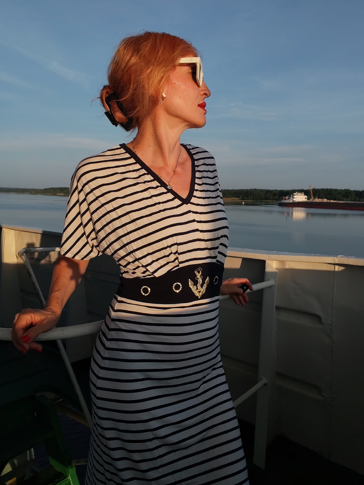 A woman with red hair pulled into a bun, wearing large white sunglasses, red lipstick, and a black and white striped dress with a black belt adorned with an anchor and two circular embellishments, stands on the deck of a boat. She is leaning on a railing and looking out at a calm body of water under a blue sky with some clouds. In the background, a large barge is visible on the water, and a tree-lined shore is in the distance. The lighting suggests it is either early morning or late afternoon. This scene is set in the city of Shchetinino, Russia.