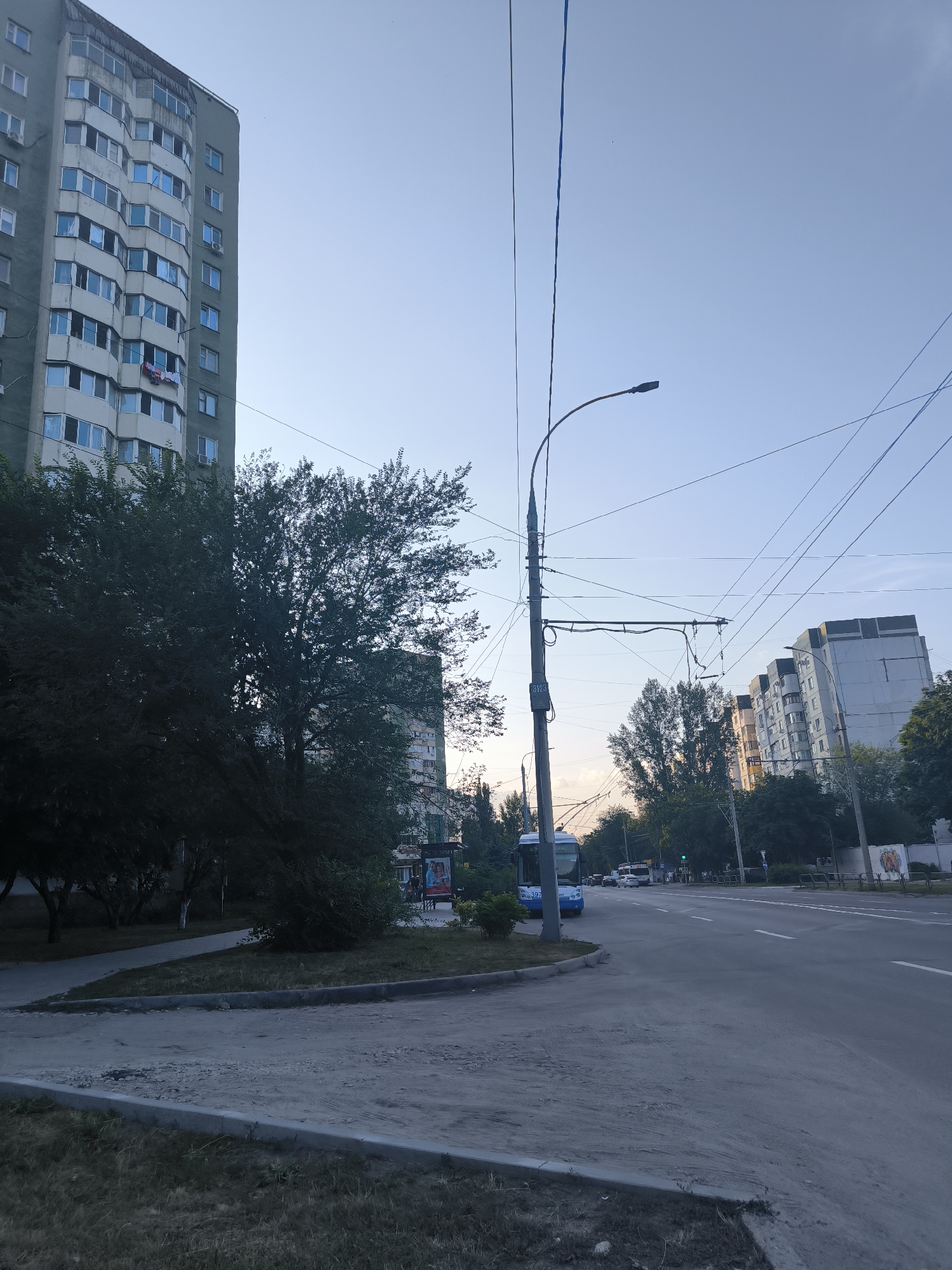 The image depicts a street scene in Chișinău, Moldova, likely during early morning or late evening given the soft lighting and clear sky. On the left side of the frame, a tall, light green apartment building with numerous windows dominates the foreground. Adjacent to it, dense green trees create a shaded area.
A multi-lane road runs through the center of the image, with a white bus visible on the far side of the road, heading away from the viewer. Overhead, a complex network of trolleybus wires crisscrosses the sky, supported by poles along the street. The road surface is asphalt, with white lane markings visible. On the right side of the road, more apartment buildings of various heights and architectural styles are visible, interspersed with trees.
The area in front of the apartment building on the left appears to be a small park or green space, with a paved walkway curving through it. The ground in the immediate foreground is covered with gravel and patchy grass, suggesting ongoing construction or maintenance. The overall impression is of a typical urban environment in a post-Soviet city.