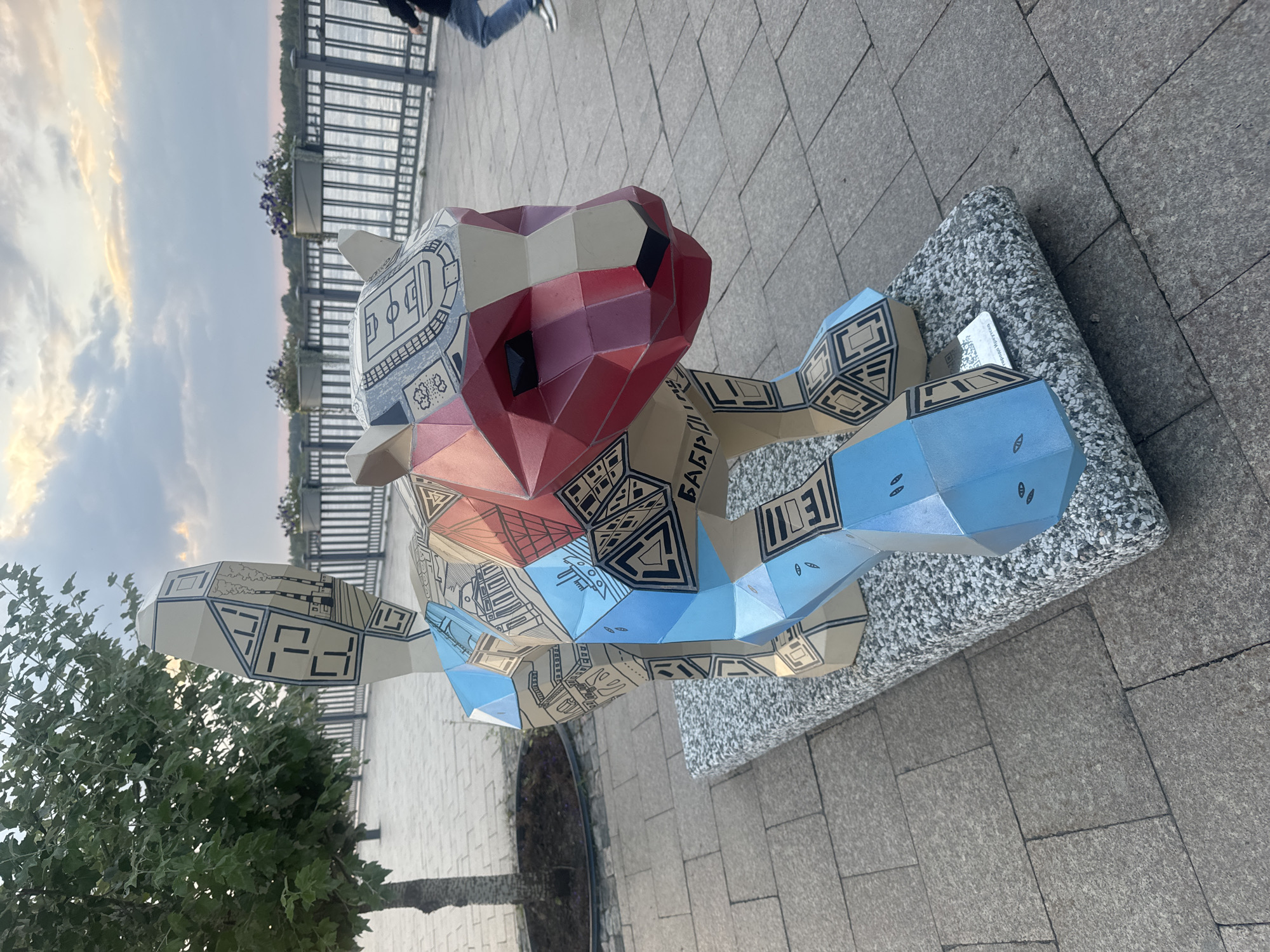 The image shows a sculpture of a polygonal animal, possibly a tiger or a lion, on a granite pedestal. The sculpture is decorated with geometric patterns and images of city landmarks, including buildings, a stadium, and a ship. The animal sculpture is primarily beige, with red on its face and blue on its legs. The background shows a paved walkway, a fence, a body of water, and some trees. In the far right of the image, a person is walking away from the camera. The sky above is cloudy with hints of sunset colors. A small sign with a QR code is visible on the pedestal. The sculpture appears to be part of a public art installation. The Cyrillic text "БАБР" is visible on the sculpture, which refers to the Siberian tiger, the symbol of Irkutsk, Russia.
