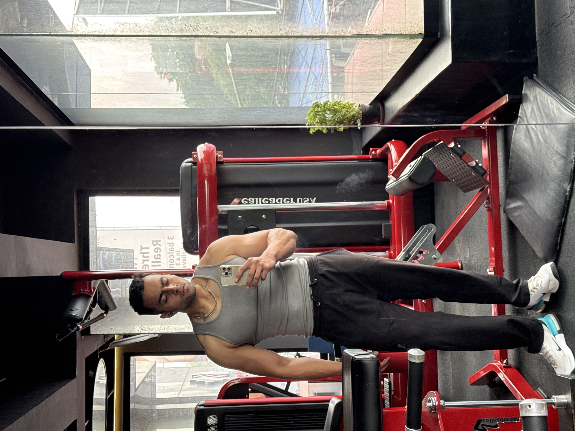 The media file displays a young man in a gym, standing next to a large piece of red and black exercise equipment. He has a muscular physique, dark hair, and is wearing a grey tank top, black track pants, and white sneakers with blue accents. A thin chain is visible around his neck. He is holding a light-colored smartphone (possibly an iPhone with multiple cameras) in his right hand, appearing to look down at its screen or posing for a selfie, with his eyes closed or cast downwards.
The setting is an indoor gym, characterized by dark walls and large windows in the background that allow natural light in. Through the windows, reflections of an urban environment are visible, showing buildings, possibly scaffolding or construction, suggesting a city location like Kolkata, India, during daytime. A small green potted plant is placed on a ledge near one of the windows.
The prominent gym equipment beside the man is large, red-framed, and features black padding and a textured footrest, likely a leg press or similar strength training machine. Partially visible, inverted text on the machine reads "ASU 139D69S", indicating a brand or model. Additional indistinct, inverted text can be seen in the window reflections, possibly from external signs, including fragments like "The Reg..." and "svalie...". The man's expression is neutral and focused on his phone, suggesting a moment of rest or preparation during a workout.