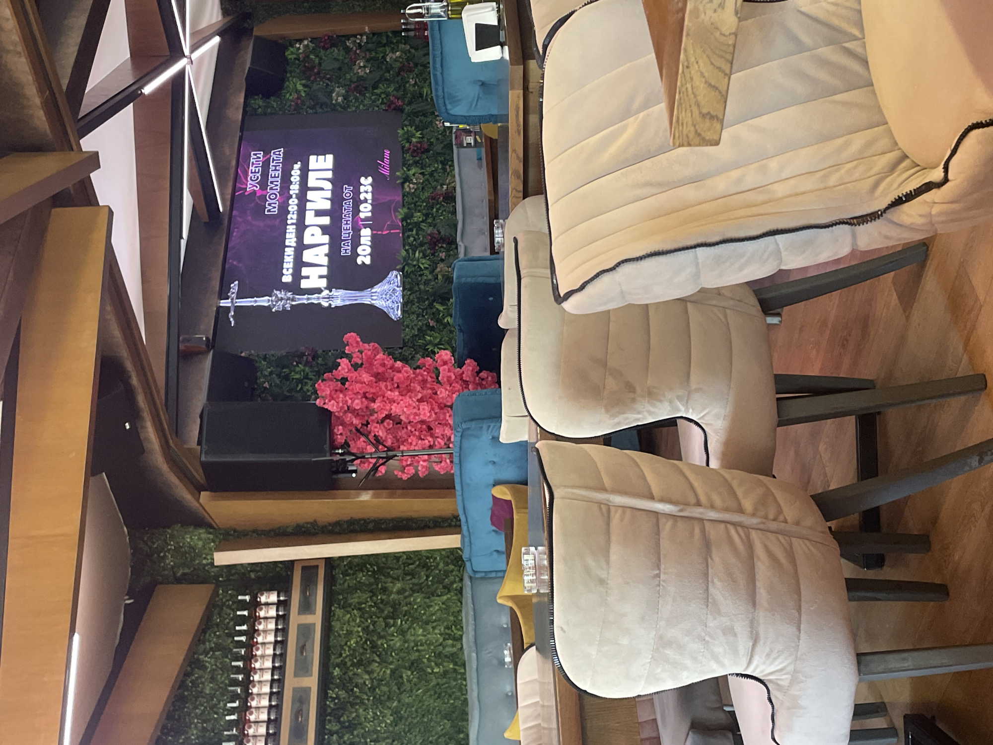 Here is a forensic description of the image:
The image depicts an indoor setting, possibly a restaurant or bar, located in Sofia, Bulgaria. The composition is focused on seating arrangements consisting of upholstered chairs with light-colored cushions and dark piping details.
A poster with the inscription "УСЕТИ МОМЕНТА" (which translates to "Feel the Moment") and the word "НАРГИЛЕ" (which translates to "Hookah") is visible, alongside the pricing "20лв 10.23€," suggesting an advertisement for hookah services at the venue, every day from 12:00-18:00. The location "Milano" is also present on the poster.
The scene features decorative elements such as greenery and pink floral arrangements, adding an element of ambiance. Shelving containing rows of bottles is also apparent. The location appears to be well-lit with both natural and artificial lighting. No individuals are visible in the frame.