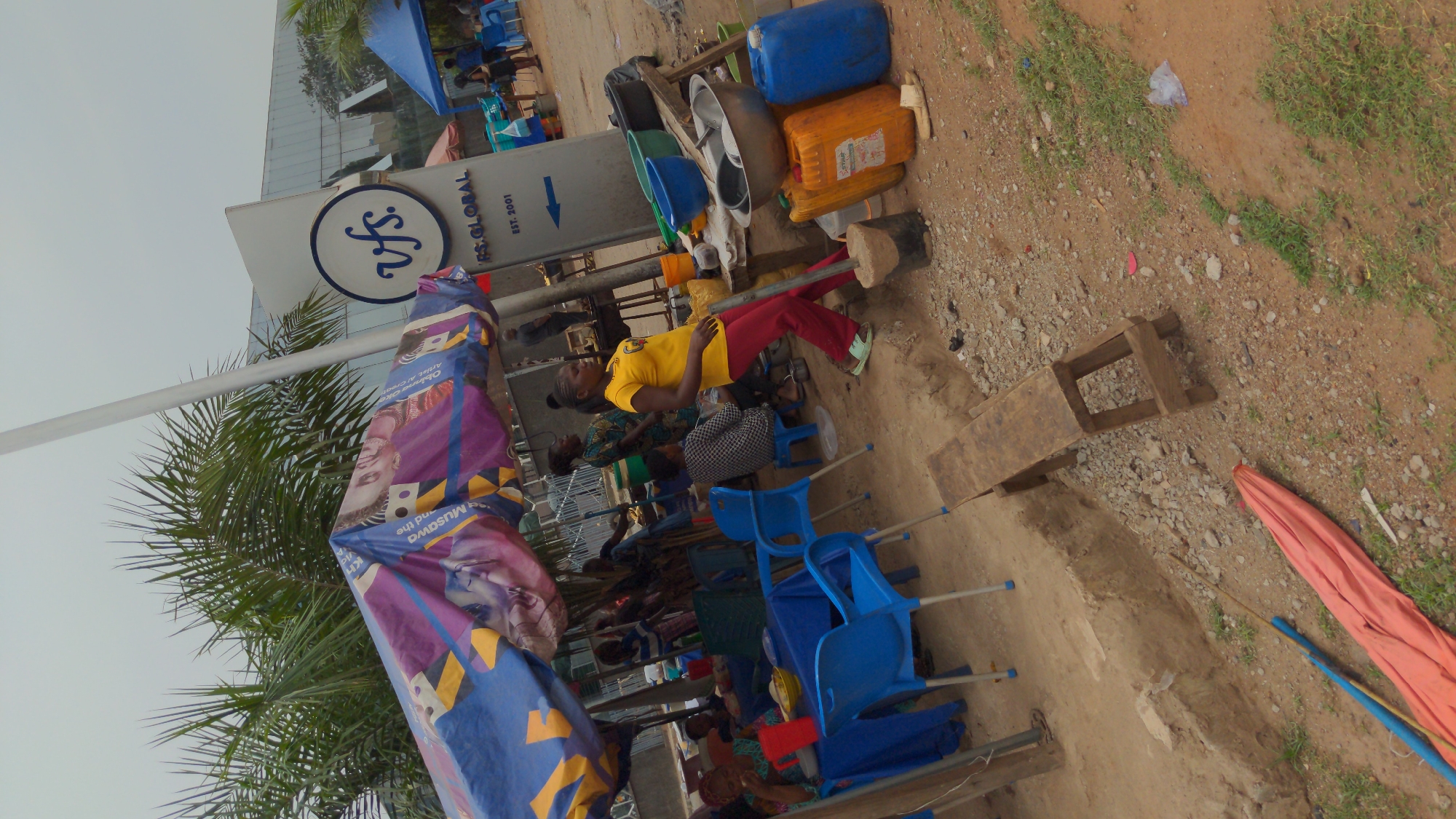 This media file captures an outdoor scene, likely a waiting area outside a service center in Abuja, Nigeria. The bright, diffuse lighting suggests daytime with an overcast sky. The ground is unpaved, composed of dirt and gravel with scattered patches of green grass.
Numerous individuals, including adults and possibly children, are present, predominantly seated on various chairs. Many are on simple blue plastic chairs, while others are on wooden stools or benches. A woman in a yellow t-shirt and red trousers is prominent, leaning against a metallic support pole. Another person, dressed in patterned clothing, sits nearby. People appear to be waiting or congregating in an informal setting.
A large, colorful canopy or banner, primarily blue and purple with yellow accents, provides shade over part of the waiting area. Behind it, a palm tree's fronds are visible, and a modern building with glass facades stands in the background.
Visible objects include several blue and orange plastic jerrycans, commonly used for liquids, and large metal basins or pots, indicating a functional outdoor environment. A long white pole stands near the central sign.
A prominent white sign with a circular blue "vfs" logo clearly displays "VFS Global" and "EST. 2001," along with an arrow. This suggests the location is a VFS Global visa application center or a similar administrative service point. The colorful banner also features partial text, including "MUSawa and the," "Kh," "Artist A/C," and "Obi," which seem to be parts of an advertisement or event promotion. The overall atmosphere conveys a routine public gathering and waiting area.