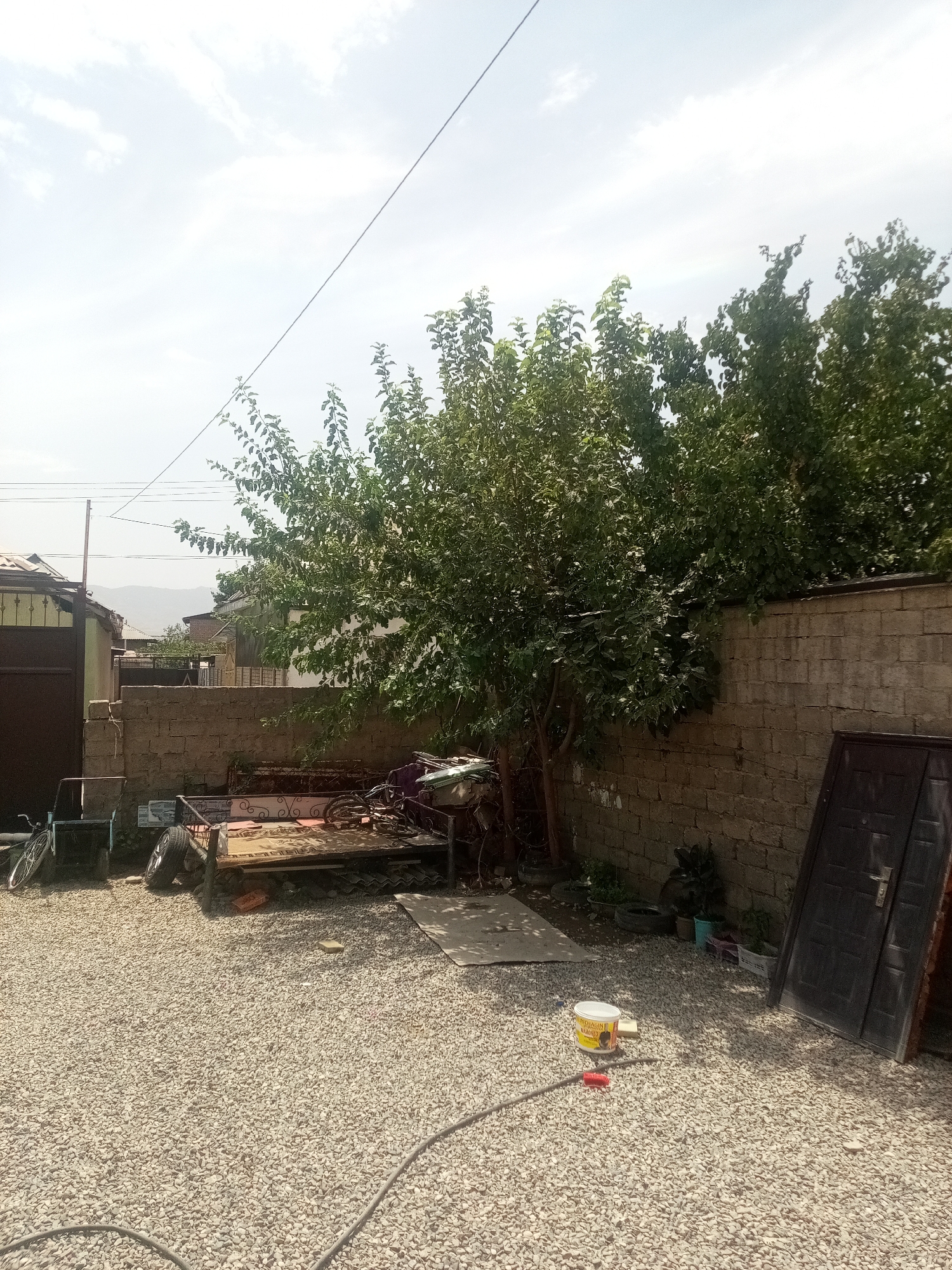 A wide shot captures a cluttered outdoor yard in an unknown city. The ground is covered in pale gravel. To the left, a dark brown metal gate is partially visible, attached to a light-colored building. In front of the gate, a metal chair and the rear wheel of a bicycle are visible.
Dominating the center of the frame is a large, leafy tree casting shadows. In front of the tree, a dilapidated metal bed frame with a patterned mattress lies on the gravel. Scattered around the bed are various objects, including a bicycle, tires, and what appears to be scrap metal. Behind the bed, a brick wall runs horizontally across the image, and on top of it, more greenery from trees can be seen. To the right of the brick wall, a dark, tall metal door stands leaning against the wall.
In the foreground, a white bucket with a yellow lid and some red tubing are placed on the gravel. In the background, several buildings and distant hills can be observed under a bright, overcast sky with thin clouds and overhead power lines. The scene suggests a storage or work area.