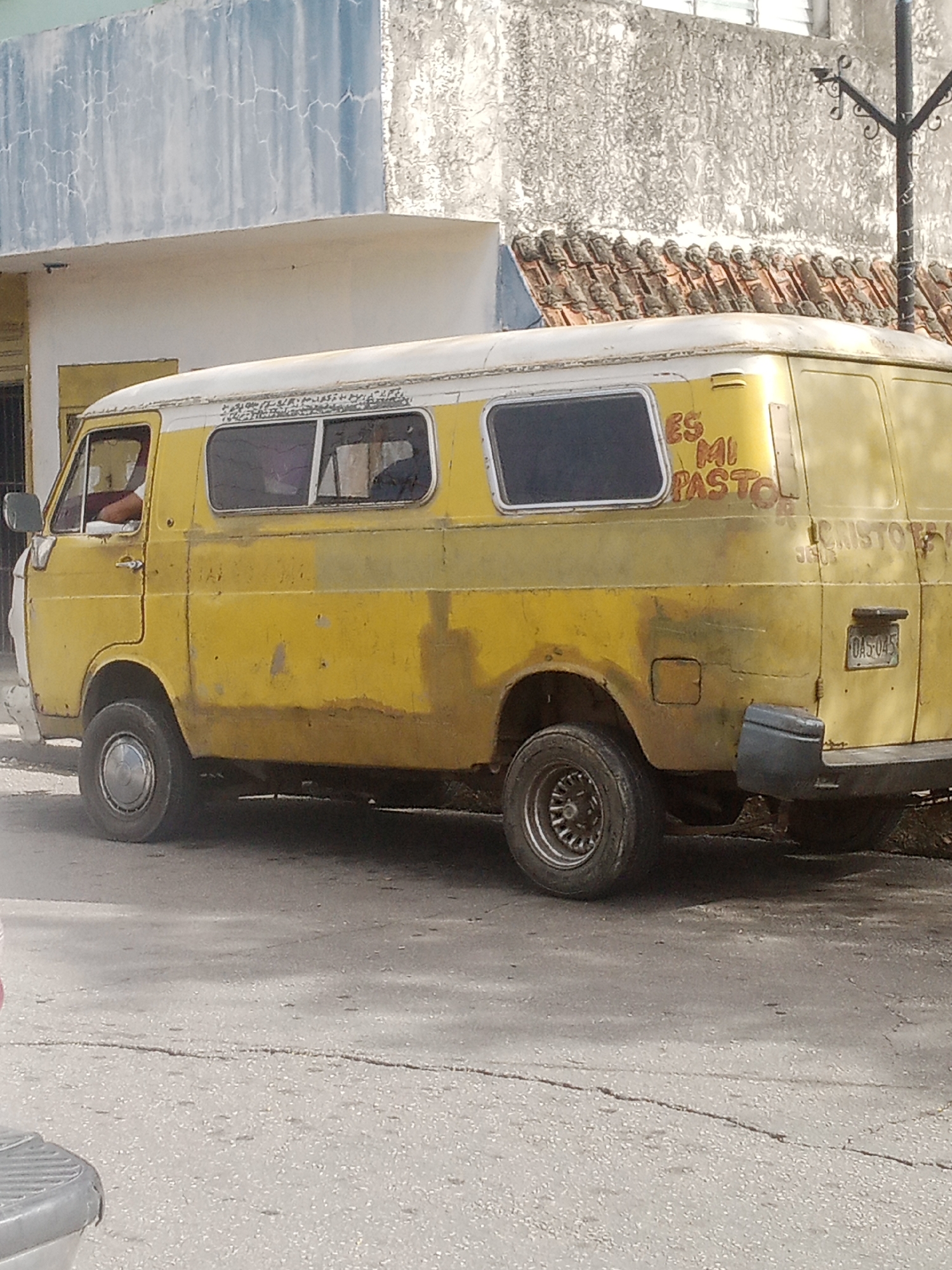 The image shows a yellow and white van parked on a cracked asphalt street in Santa Ana, Venezuela. The van has a white roof and yellow body, with some rust and dirt visible. There is writing on the side of the van that reads "ES MI PASTOR" and "CRISTO", indicating a religious message. The van has two side windows and a rear window. A license plate on the rear of the van is visible with the characters "OA5-045". The background features a building with a light blue and white facade and a terracotta tiled roof. A street lamp with an ornate black metal pole is also visible. The overall scene suggests a typical street view in a Venezuelan town.