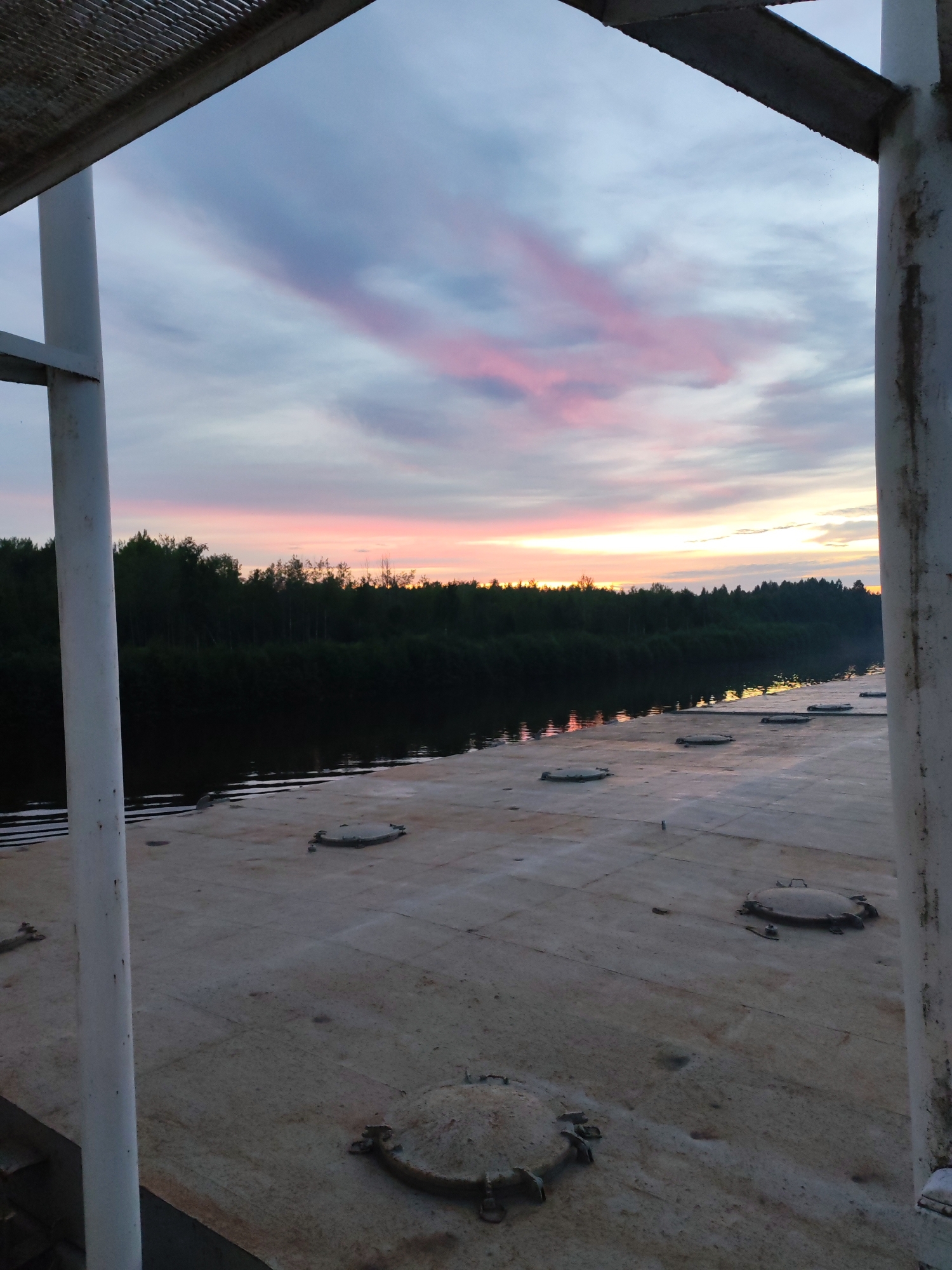 The image depicts a twilight scene overlooking a body of water and a treeline. The sky displays a gradient of colors, from pale blue at the top to shades of pink and orange near the horizon. There are wispy clouds streaked with pink. The water is dark and reflects the sky's colors. A dense forest forms a dark silhouette along the opposite bank. In the foreground, a flat, textured surface, possibly a deck, is visible. This surface has several circular, metal hatches, some appearing weathered and rusty. White vertical poles and a metallic grate are also present, framing the view. The overall impression is of a tranquil, natural landscape at dusk, viewed from a man-made structure like a boat or pier. The location is specified as Kurdyug, Russia. There are no people or animals visible in the image.