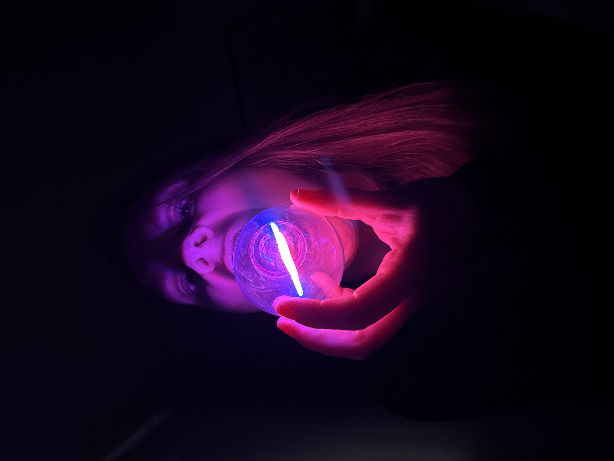 A person with long, light-colored hair is holding a glowing orb in their right hand. The orb emits blue and purple light and is held up close to the person's face. The person is lying down, facing upwards. Their face is partially illuminated by the orb's light, revealing their eyes and nose. The background is dark. The location is unknown and cannot be determined from the image.