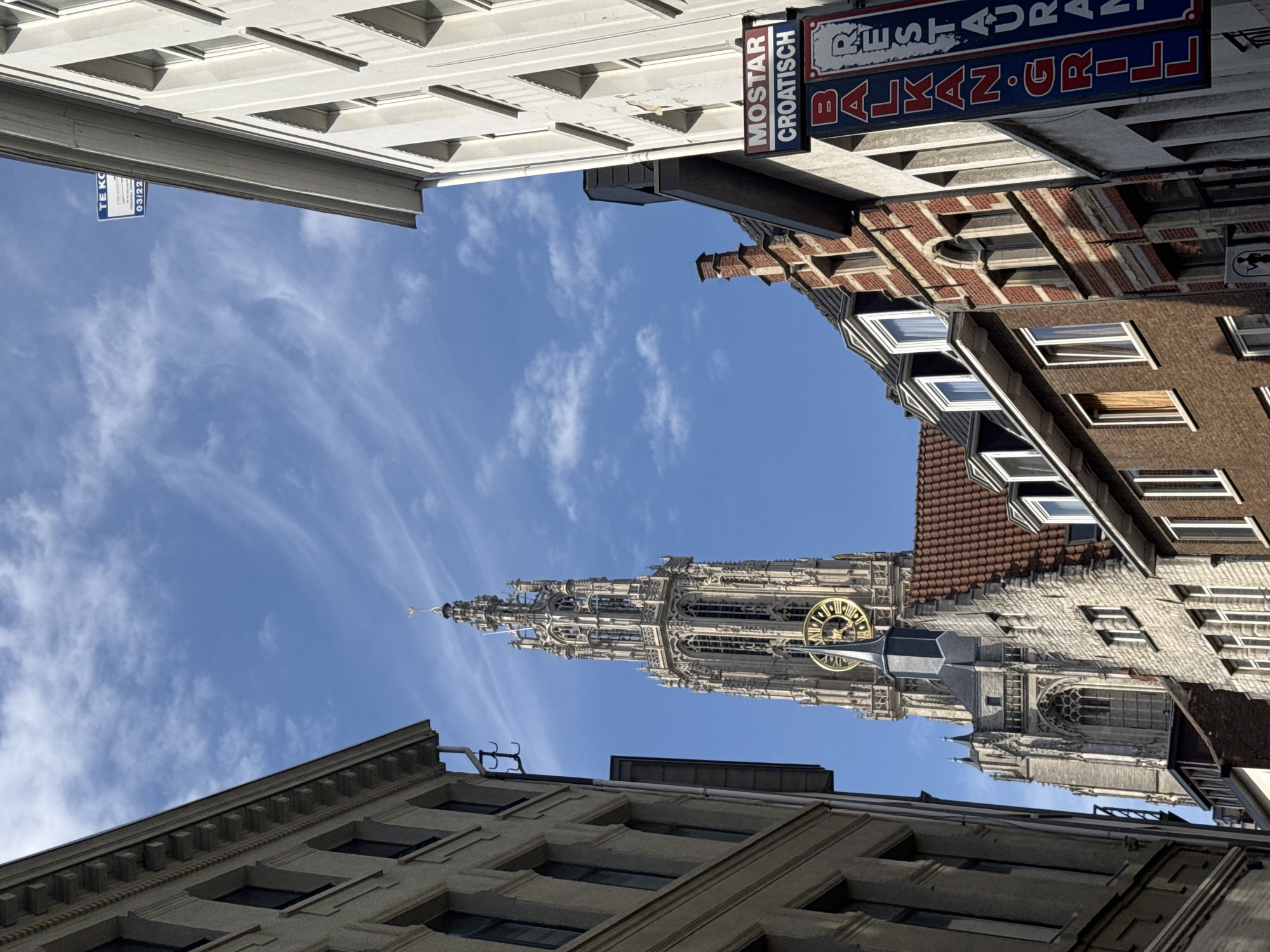 The image captures a street view in Antwerp, Belgium, dominated by the towering spire of the Cathedral of Our Lady. The cathedral's intricate Gothic architecture, including its clock face, is prominently featured against a bright blue sky with wispy white clouds. The foreground is flanked by buildings on either side, suggesting a narrow street. On the right, a building displays a sign for "MOSTAR CROATISCH" and "BALKAN GRILL RESTAURANT," indicating dining establishments. Another sign is visible higher up on the right-side building with Dutch text. The left side of the image shows the facade of a building with arched windows. The overall scene depicts an urban environment with historical architecture and commercial activity.
