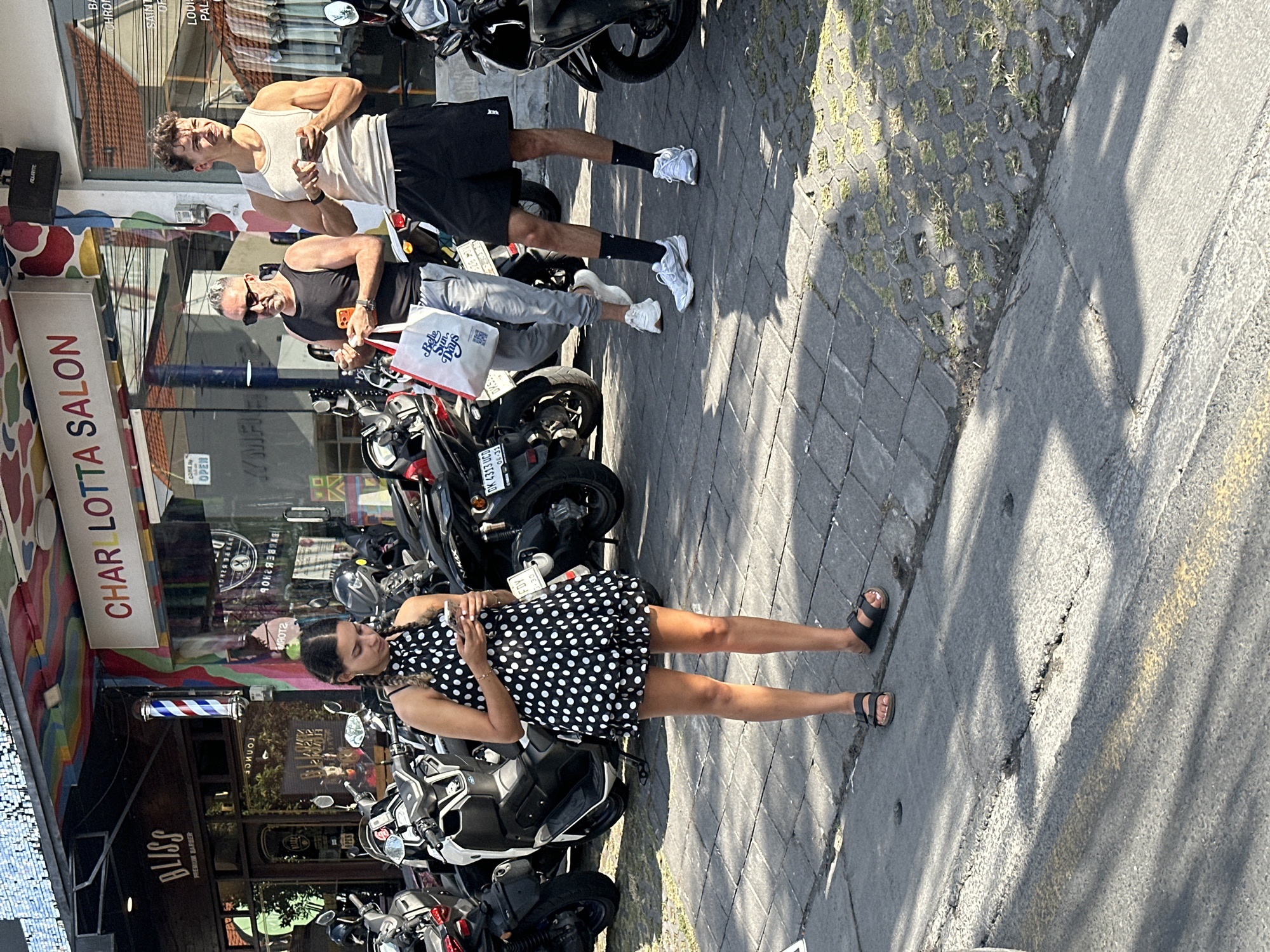 In North Kuta, Indonesia, on a sun-drenched street, a young woman with braided hair stands facing away from the camera, engrossed in her phone. She wears a short, black and white polka dot dress and black sandals. Behind her, two men stand on the sidewalk. The man on the left, wearing a black tank top and sunglasses, holds an orange-colored phone and a white reusable shopping bag. The man on the right, in a white tank top and black shorts, also holds a phone. Parked scooters line the street in front of a building with colorful murals and signage that reads "CHARLOTTA SALON." The ground is paved with interlocking bricks and asphalt, with some patches of grass growing between the bricks. Shadows cast by the building and people stretch across the pavement.