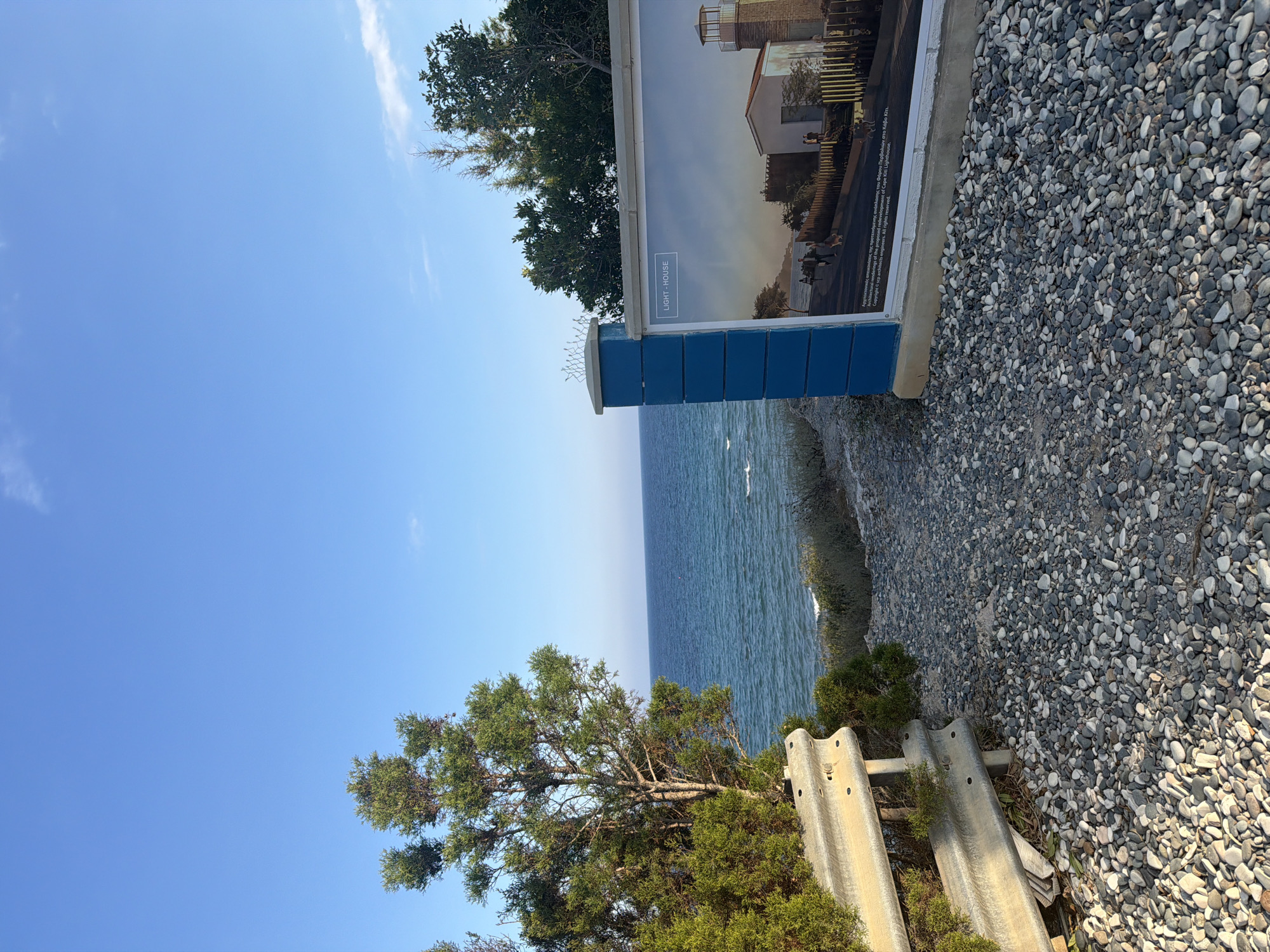 The image shows a clear blue sky with a few wispy clouds over a tranquil blue sea. In the foreground, a gravel path leads towards the sea, bordered on the left by a metal guardrail and lush green foliage. To the right, a blue-painted wooden post supports a large, horizontal sign. The sign displays an architectural rendering of a lighthouse and associated buildings, labeled "LIGHT-HOUSE". Text in both Greek and English is visible at the bottom of the sign, mentioning "proposed redevelopment of Cape Kiti Lighthouse." The scene appears to be a coastal area, likely in or near Pervolia, Cyprus, given the mention of Cape Kiti Lighthouse.