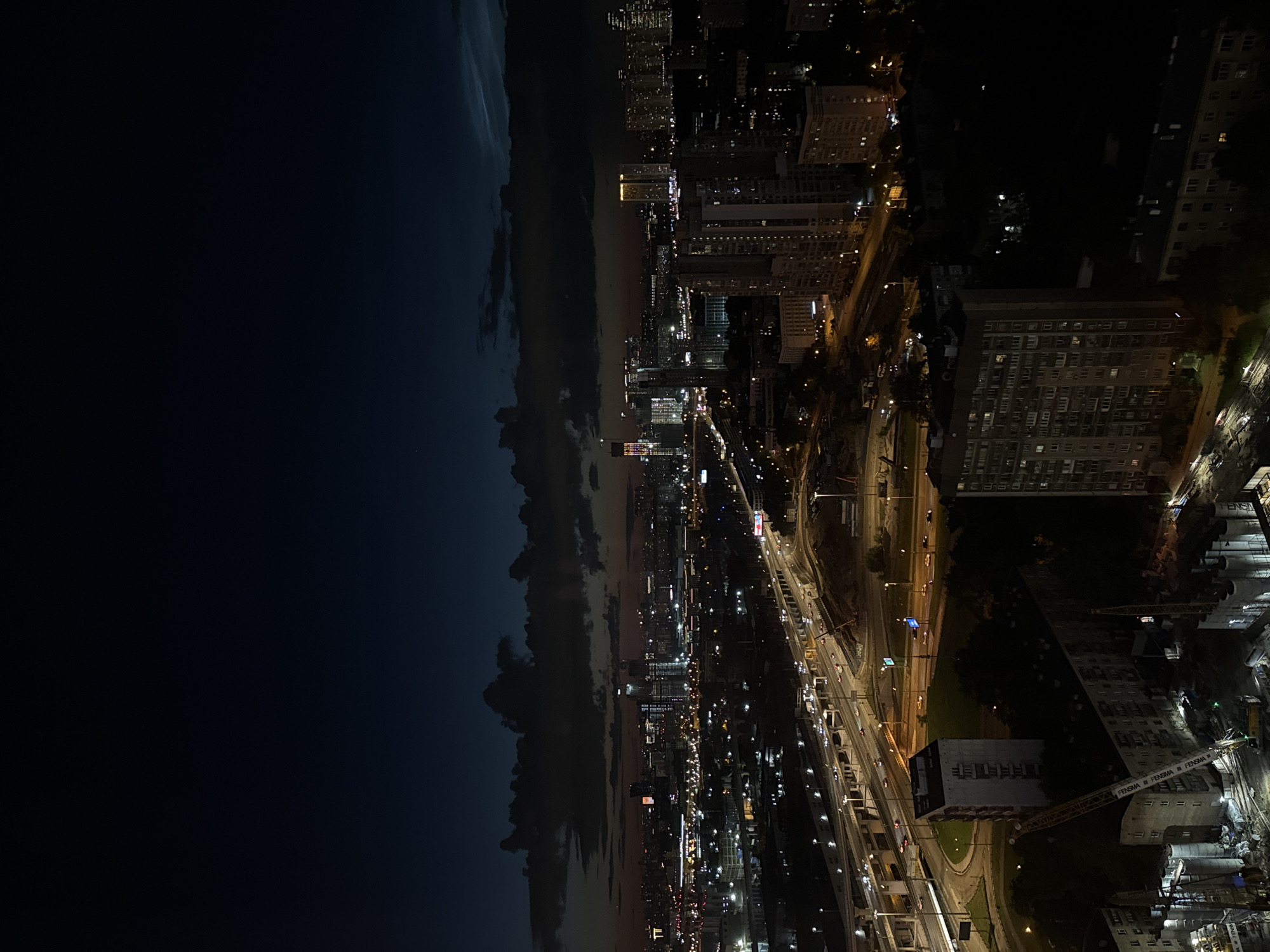 This is a high-angle, wide shot of a city skyline at dusk. The sky is a deep blue transitioning to dark blue towards the top, with scattered clouds. The city lights are visible, creating a glow across the landscape. A multi-lane highway with traffic is prominent in the mid-ground, with cars illuminated by streetlights and headlights. Several buildings of varying heights are scattered throughout the scene, including apartment buildings and taller, modern structures in the distance. There is evidence of construction with cranes and building materials visible in the foreground. The scene appears to be from the city of Khimki, Russia, given the architectural style and the presence of infrastructure consistent with the region. No people are clearly identifiable in the image. The overall atmosphere is one of urban activity at the end of the day.