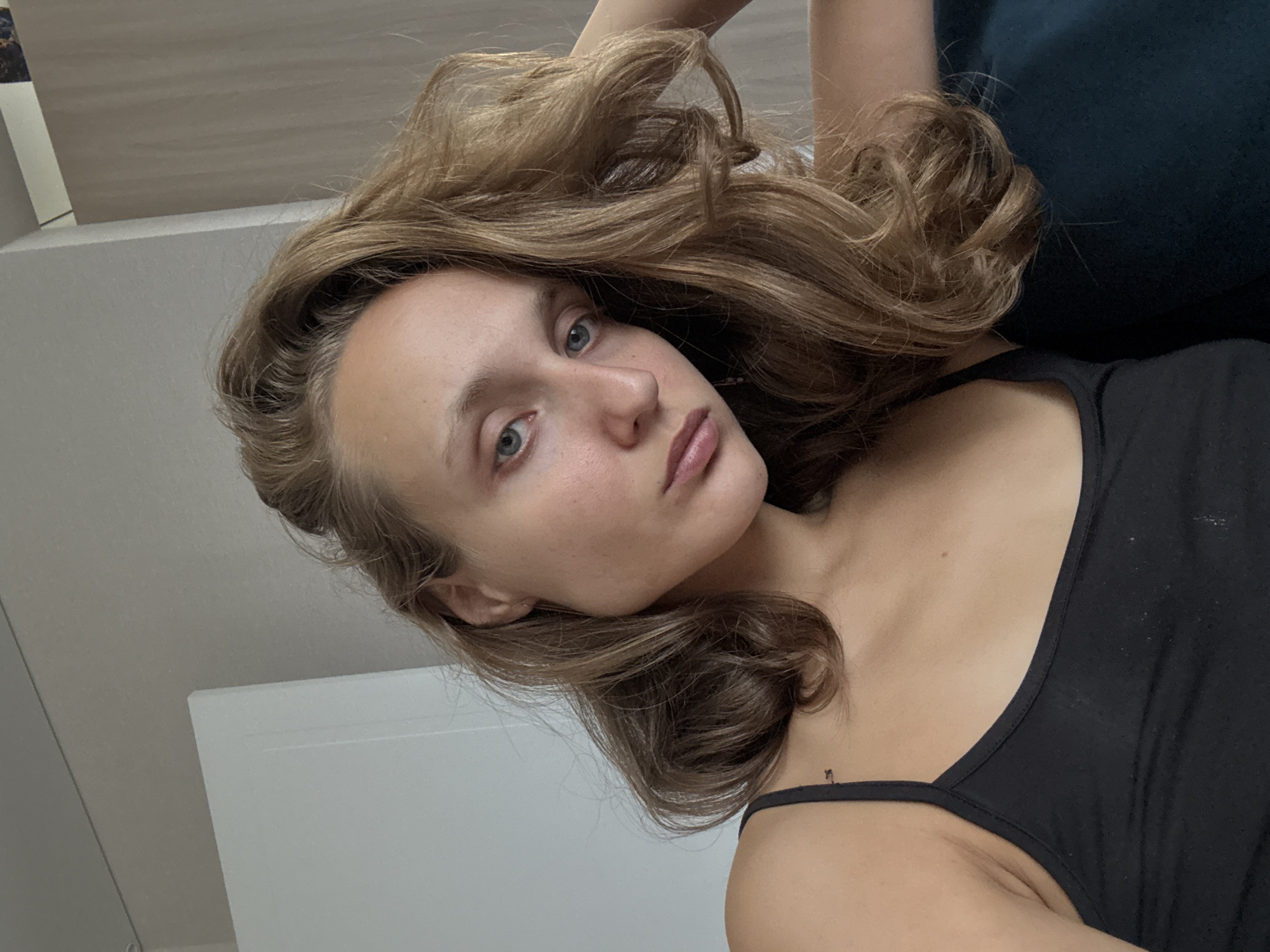 A young woman with long, wavy, light brown hair is captured in a close-up, self-taken photograph. She has blue eyes and fair skin, with her left arm resting behind her head and her right hand out of frame. She is wearing a black tank top. The background appears to be an indoor domestic setting, with a white door or cabinet on the left and a neutral-colored wall on the right. There is no indication of the location being in the city of Подшивалово, Russia.