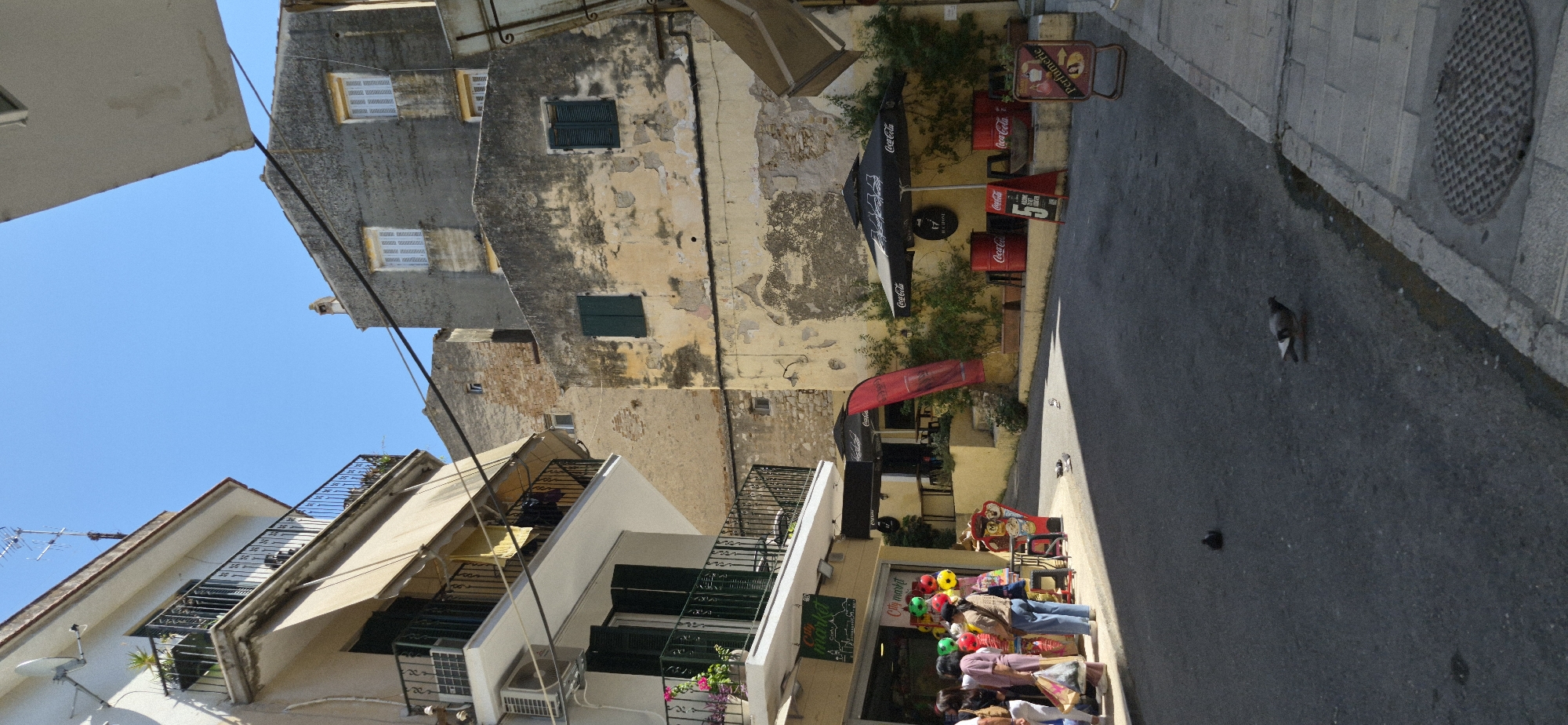The image captures a narrow, sun-drenched street in Corfu, Greece, with buildings on either side. On the left, a white building features multiple balconies with ornate black railings, an awning, and an air conditioning unit. Below, a shop with a green sign reading "City market" is visible, with several people browsing outside. Colorful balloons are displayed in front of the shop.

The right side of the street is dominated by older, weathered buildings with peeling yellow and grey plaster. Several windows with white shutters are present on these structures. A cafe or restaurant area is set up with black umbrellas bearing the Coca-Cola logo, several red Coca-Cola barrels used as tables, and outdoor seating. A red, curved flag advertising something is also visible.

The street itself is paved with asphalt, and a stone sidewalk runs along the right side, featuring a manhole cover. Pigeons are scattered across the street, some foraging, and one is in mid-stride. The sky is a clear, bright blue, indicating a sunny day. The overall impression is of a lively, warm urban scene typical of the Mediterranean.