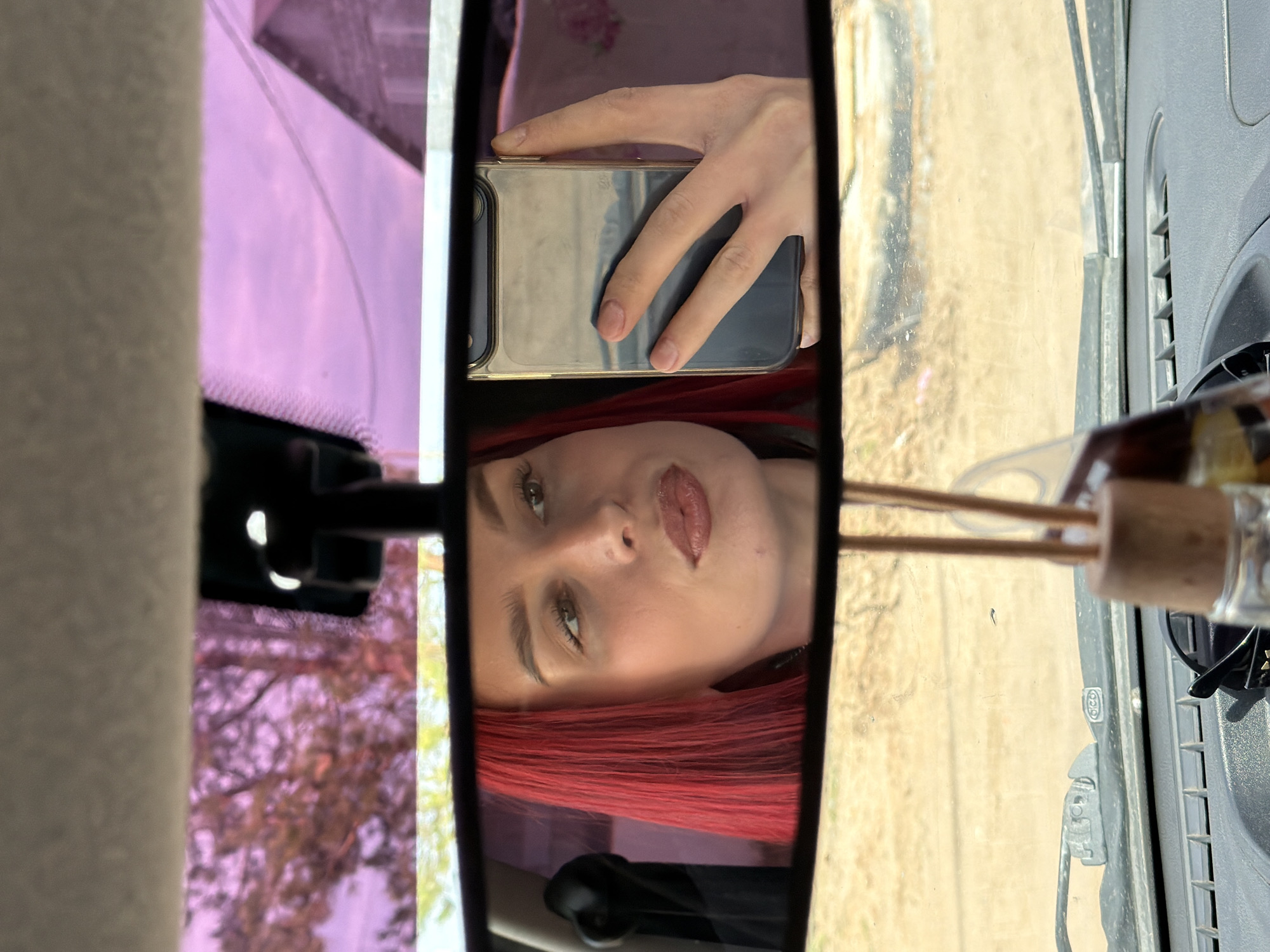 The image is a selfie taken in a car mirror, reflecting a woman with red hair holding a phone. The background includes a purple-tinted window and the dashboard of a vehicle. The reflection appears to be in a car's rearview mirror. The location in the city of Chaikovsky, Russia cannot be determined from the provided image.