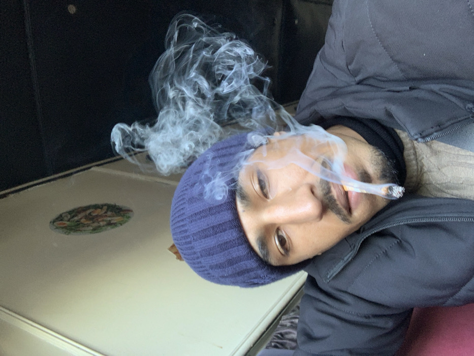 A man with a dark complexion, wearing a navy beanie and a dark puffy jacket, is angled towards the camera while smoking. Smoke curls upwards from the lit cigarette in his mouth. His beard is neatly trimmed, and he gazes at the viewer with brown eyes. The scene is lit indoors, with a white table to his left featuring an oval-shaped decorative image. A dark, possibly black, background provides a contrasting backdrop.