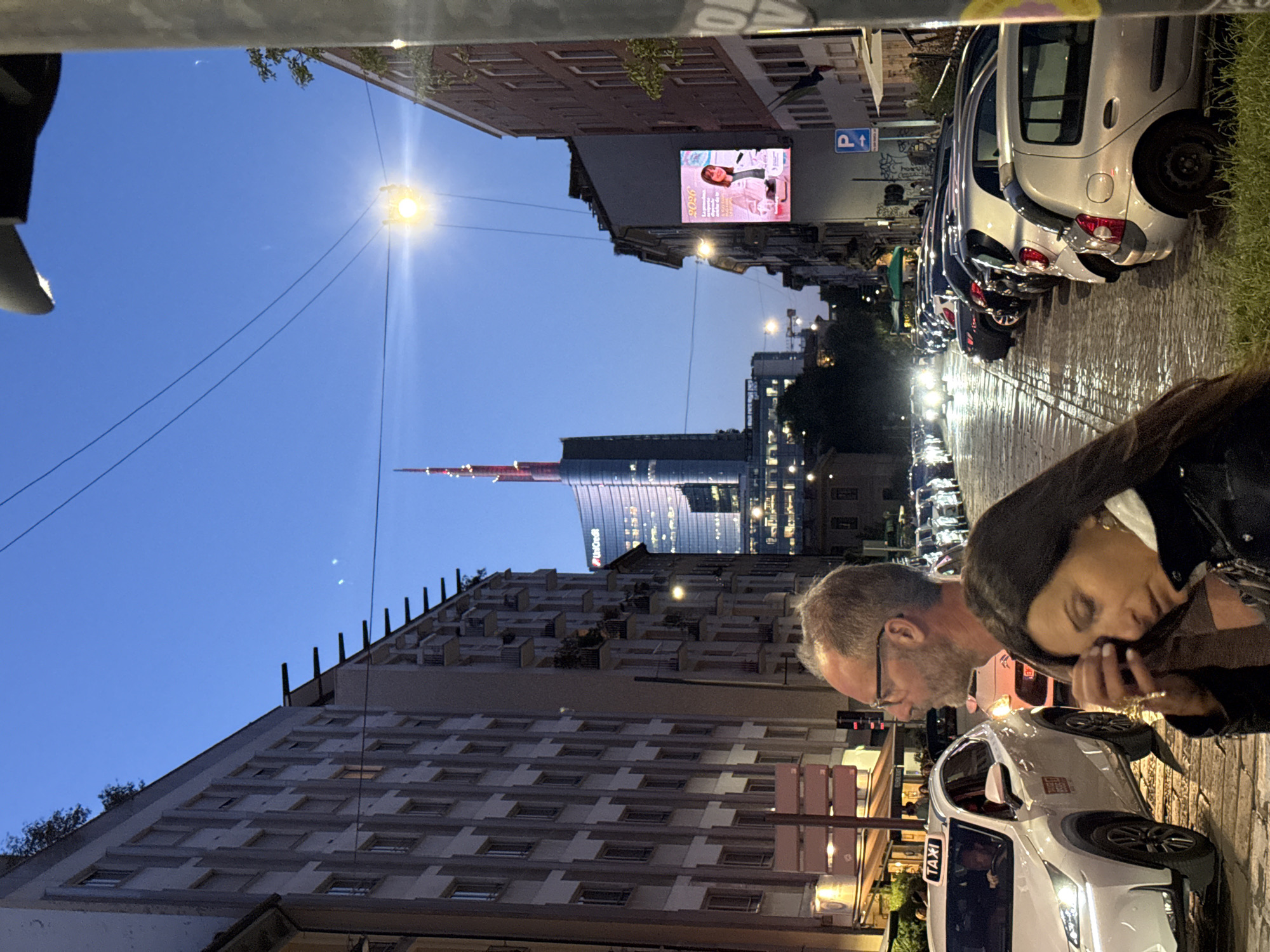 This image captures an urban street scene in Milan, Italy, at dusk or early night. The sky is a deep blue, contrasting with the numerous artificial lights illuminating the street and buildings.
In the background, the prominent Unicredit Tower, a modern skyscraper known for its red-tipped spire, stands tall, with the word "Unicredit" visible on its facade. The street below appears wet and reflective, mirroring the glow of car headlights, taillights, and streetlights, suggesting recent rain.
Buildings line both sides of the street. On the left, a digital billboard displays an advertisement featuring a woman, with "2026" and partial Italian text "La persona che si prende cura di sè stessa..." (The person who takes care of herself...) clearly visible. A blue "P" sign for parking is also present on this side.
In the foreground on the right, two people are present. A man with a beard and glasses is looking downwards. Beside him, a woman with dark hair is also looking down, her face partially obscured, possibly engaged with an object in her hands. They appear to be standing on the sidewalk. Nearby, a white taxi is visible with "TAXI" clearly labeled on its roof light, suggesting activity related to transportation. A line of parked cars extends along the street. Overhead wires crisscross the frame against the darkening sky.