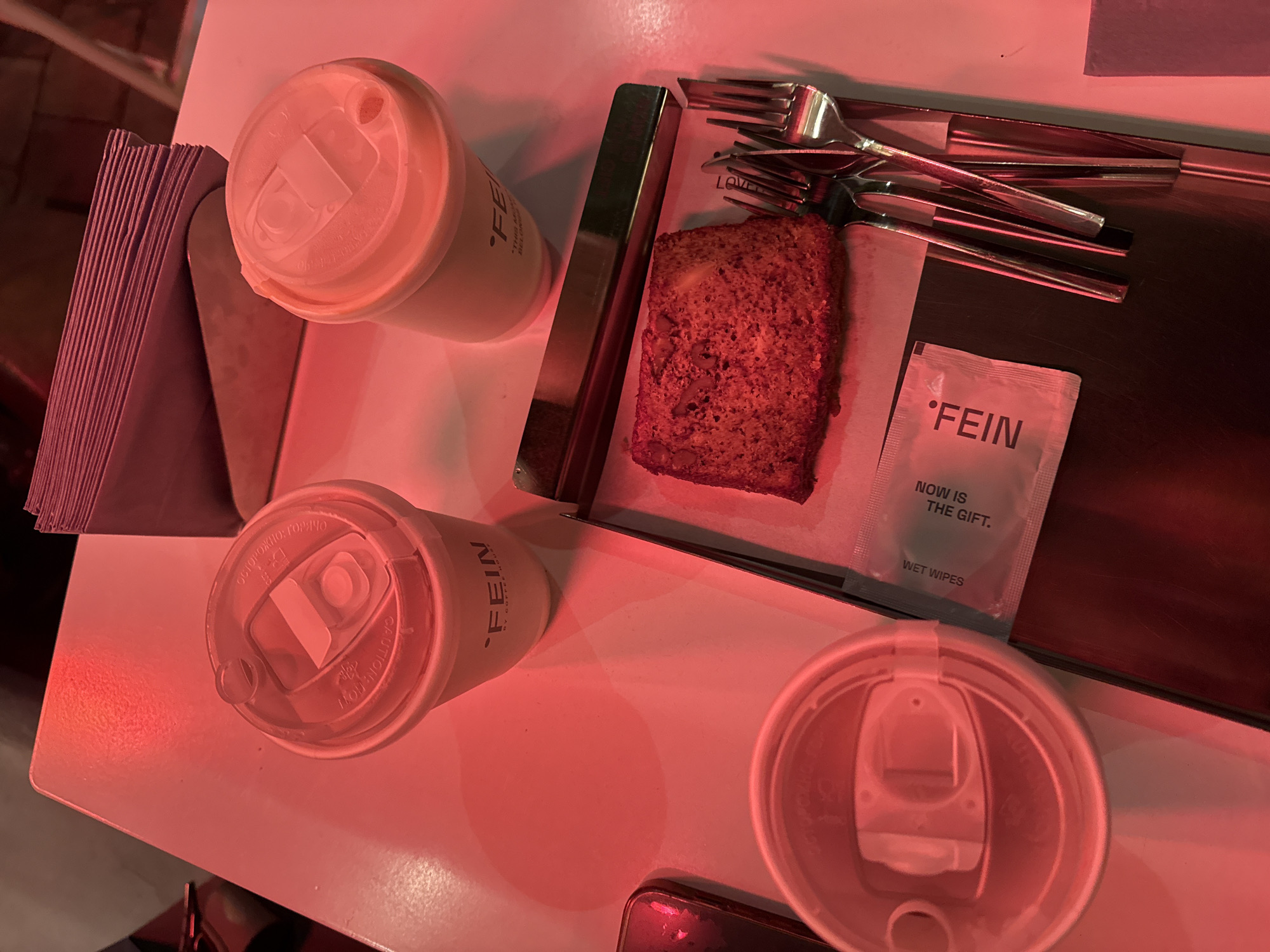 The image presents a top-down view of a table setting, likely in Yerevan, Armenia, based on the branding ("FEIN") present on the items. The table is predominantly white and bathed in a red light. There are three takeaway coffee cups with lids, two with the "FEIN" logo visible on their sides. A stack of napkins is located on the upper left of the frame. On a rectangular metal tray, there is a slice of cake or bread, several forks, and a sealed wet wipe packet, also branded with "FEIN" and the phrase "NOW IS THE GIFT." A mobile phone is visible on the bottom right of the frame. There are no people visible in the image.
