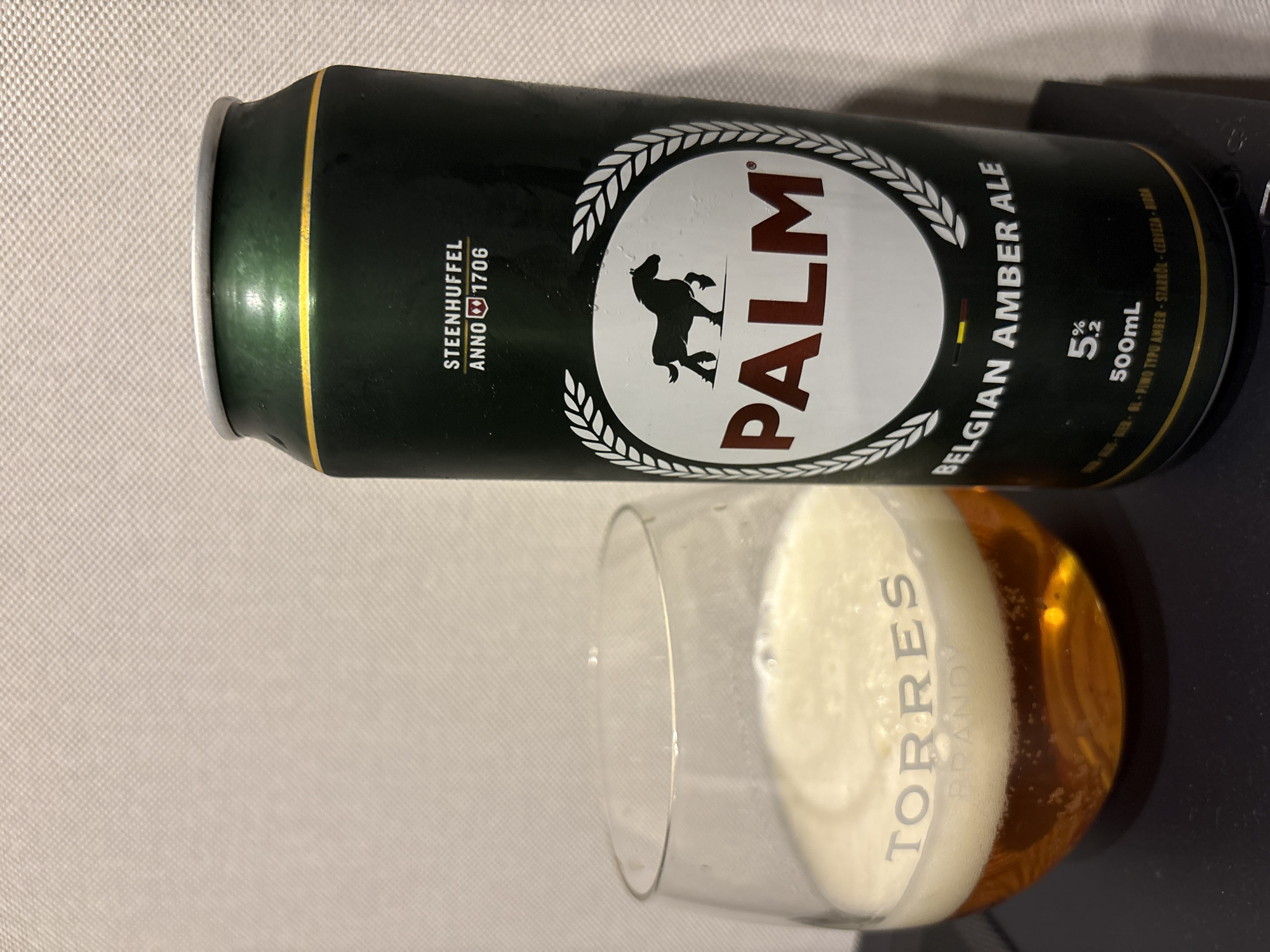 A glass of amber ale with a frothy head sits in the foreground, its rim etched with the word "TORRES". Next to it, a dark green can of Palm Belgian Amber Ale stands upright. The can features a white circular emblem with a black silhouette of a horse, and the text "PALM" in red. Above the emblem, the text "STEENHUFFEL ANNO 1706" is visible. The background is a textured, light-colored surface. The scene is indoors, with artificial lighting.
