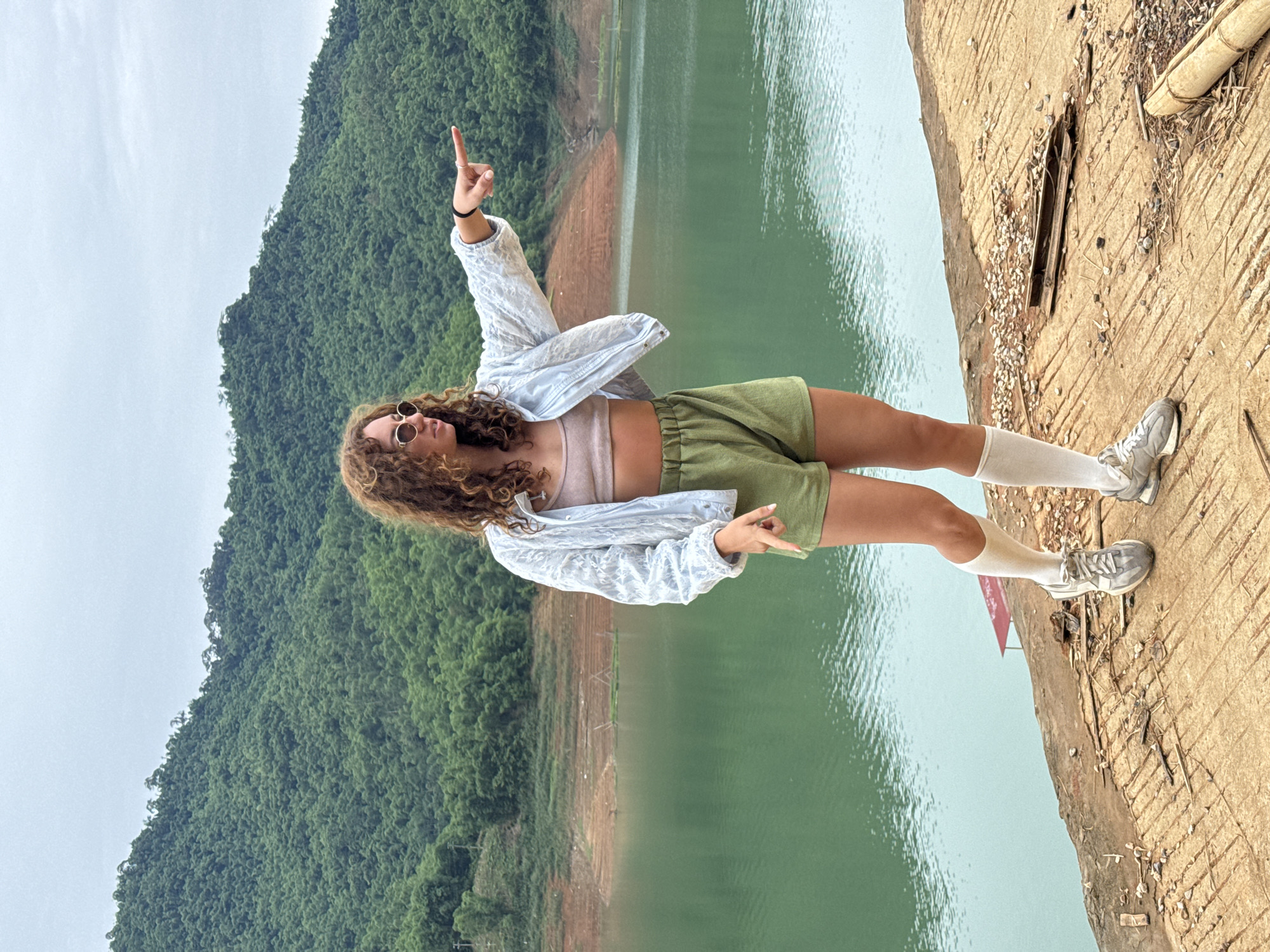 A young woman with curly brown hair stands on a concrete walkway next to a large body of green water. She is wearing round sunglasses, a light blue textured jacket, a tan crop top, olive green shorts, white socks, and gray sneakers. She is pointing to her right with her right hand and her left hand is pointing down. In the background, a lush green, tree-covered mountain rises from the other side of the water. The sky is overcast. This scene is located in Bản Phúc, Vietnam.
