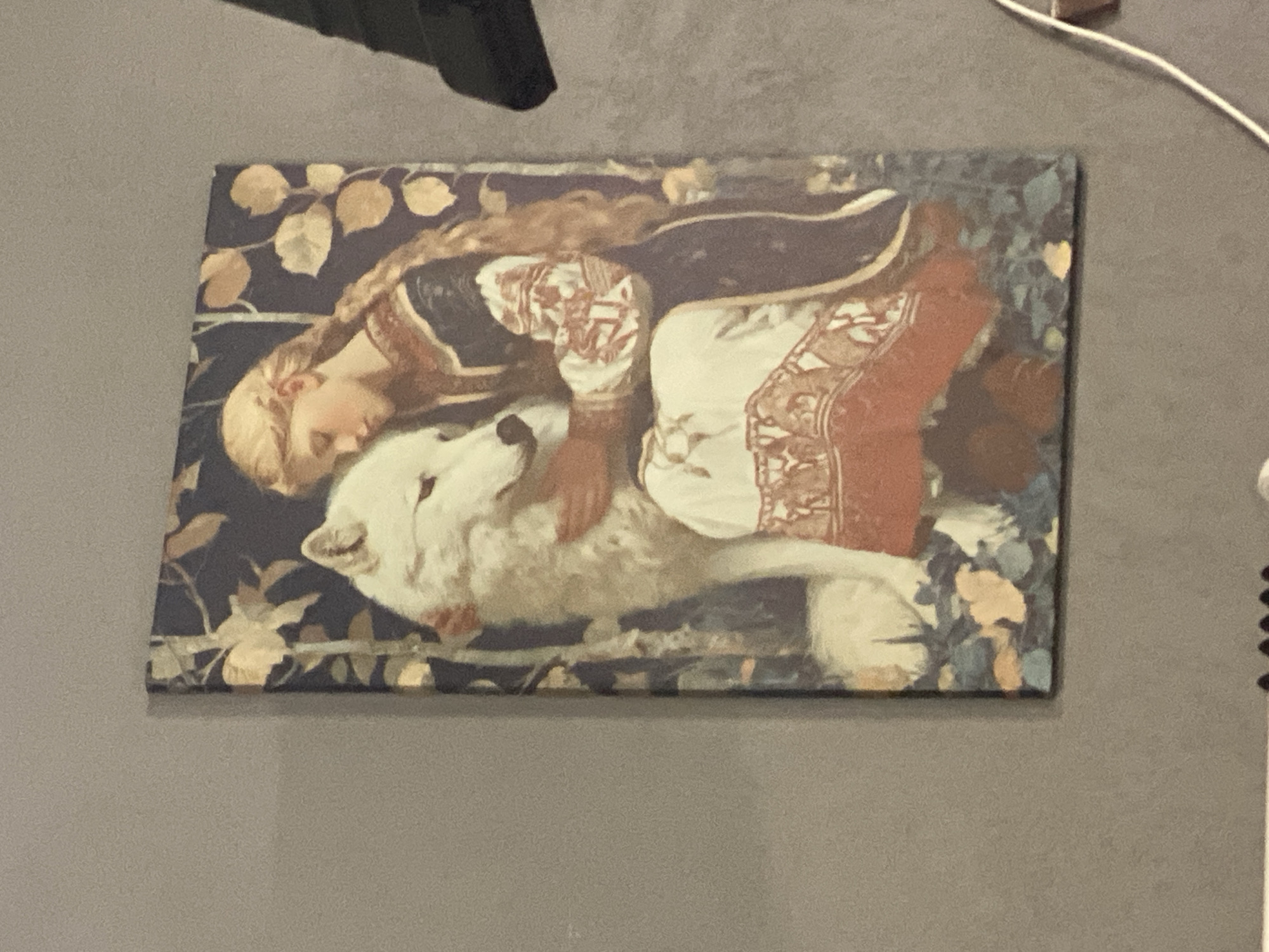 The image shows a painting of a young woman with blonde braided hair embracing a white wolf. The woman is wearing a dark blue garment with ornate red and white trim on the sleeves and a white skirt with similar trim. She is leaning her head against the wolf's head, with her eyes closed. The wolf is facing the viewer, with its head tilted slightly towards the woman. The background of the painting depicts a dark forest with autumnal leaves. The painting is displayed on a gray wall. The setting is indoors.