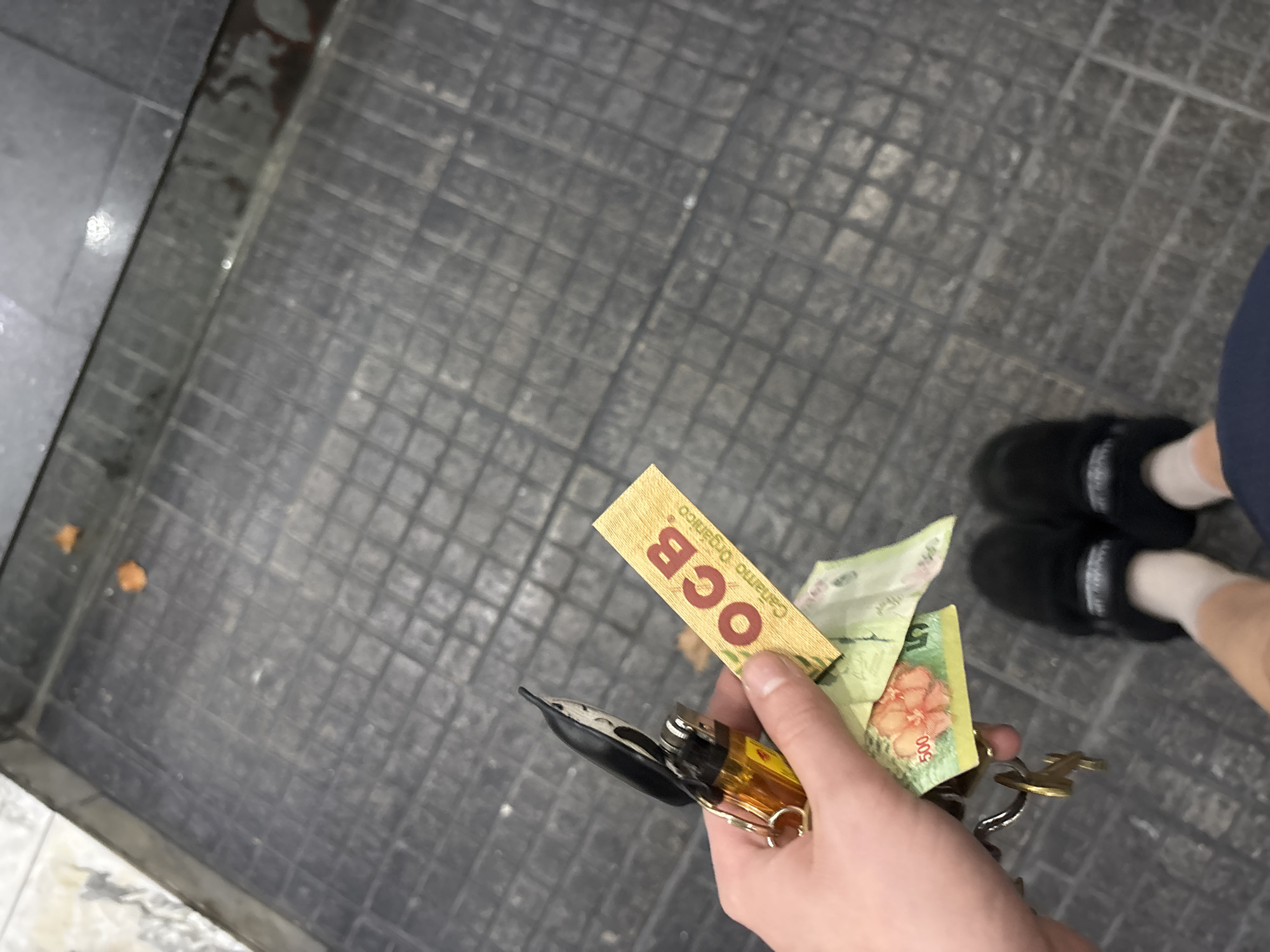 A first-person view shows a hand holding keys, Argentine currency notes, a lighter, and OCB rolling papers. The background is a gray tiled floor, possibly a public walkway. A person wearing black slippers and white socks is partially visible in the upper right corner. Based on the currency and the brand of rolling papers being OCB, the location is likely in the city of Buenos Aires, Argentina.