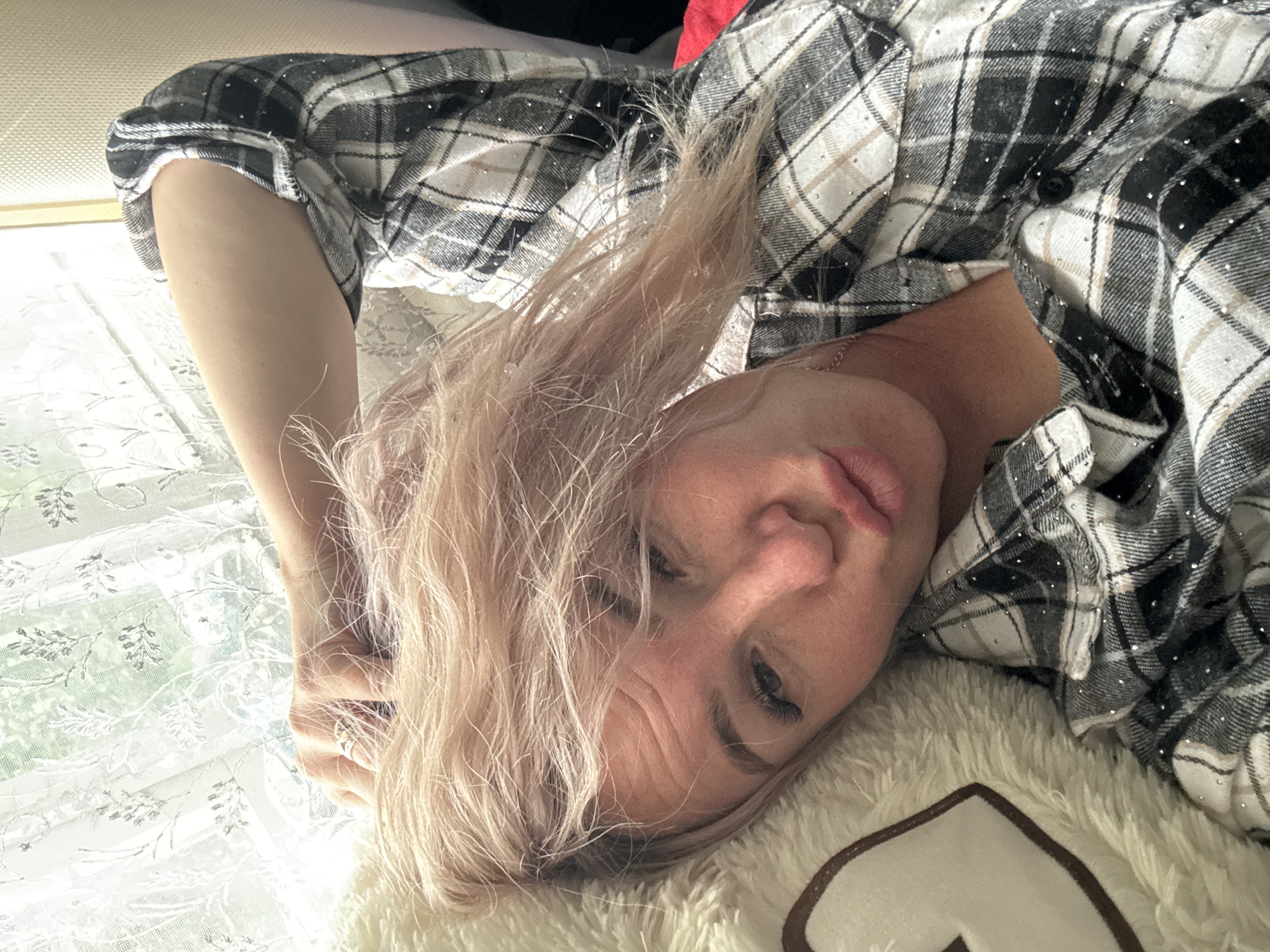A woman with light brown hair and a plaid shirt is lying down, resting her head on a fluffy pillow. Her arm is behind her head, and her hand is touching her hair. She is looking at the camera with her lips pursed. The background is a window with white curtains and a hint of greenery outside. The setting appears to be indoors, possibly a bedroom, and the lighting suggests it is daytime. There is no indication of the location being in Pozdyshki, Russia.